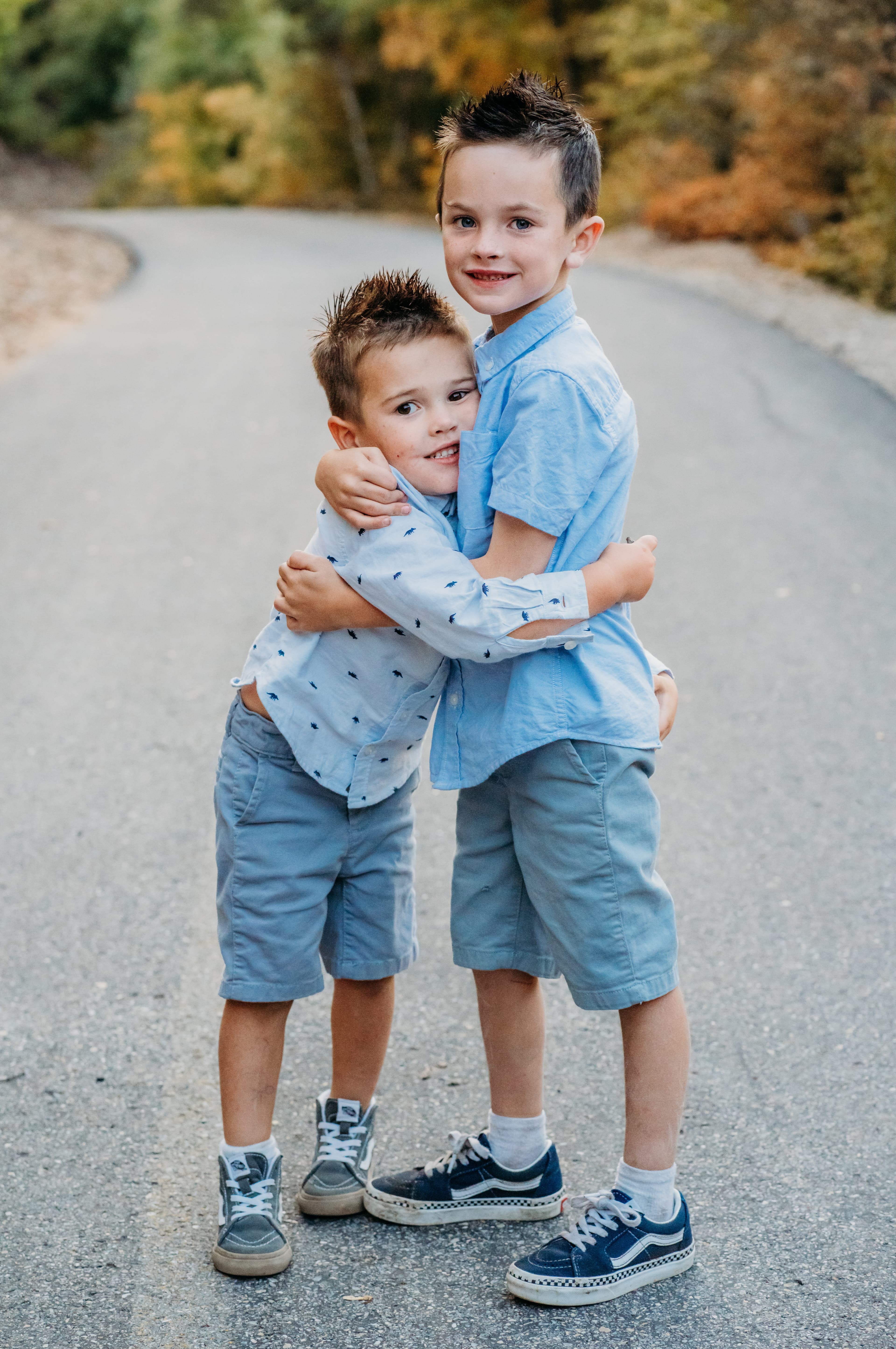 Two young brothers in matching blue shirts hug each other on a tree-lined autumn road