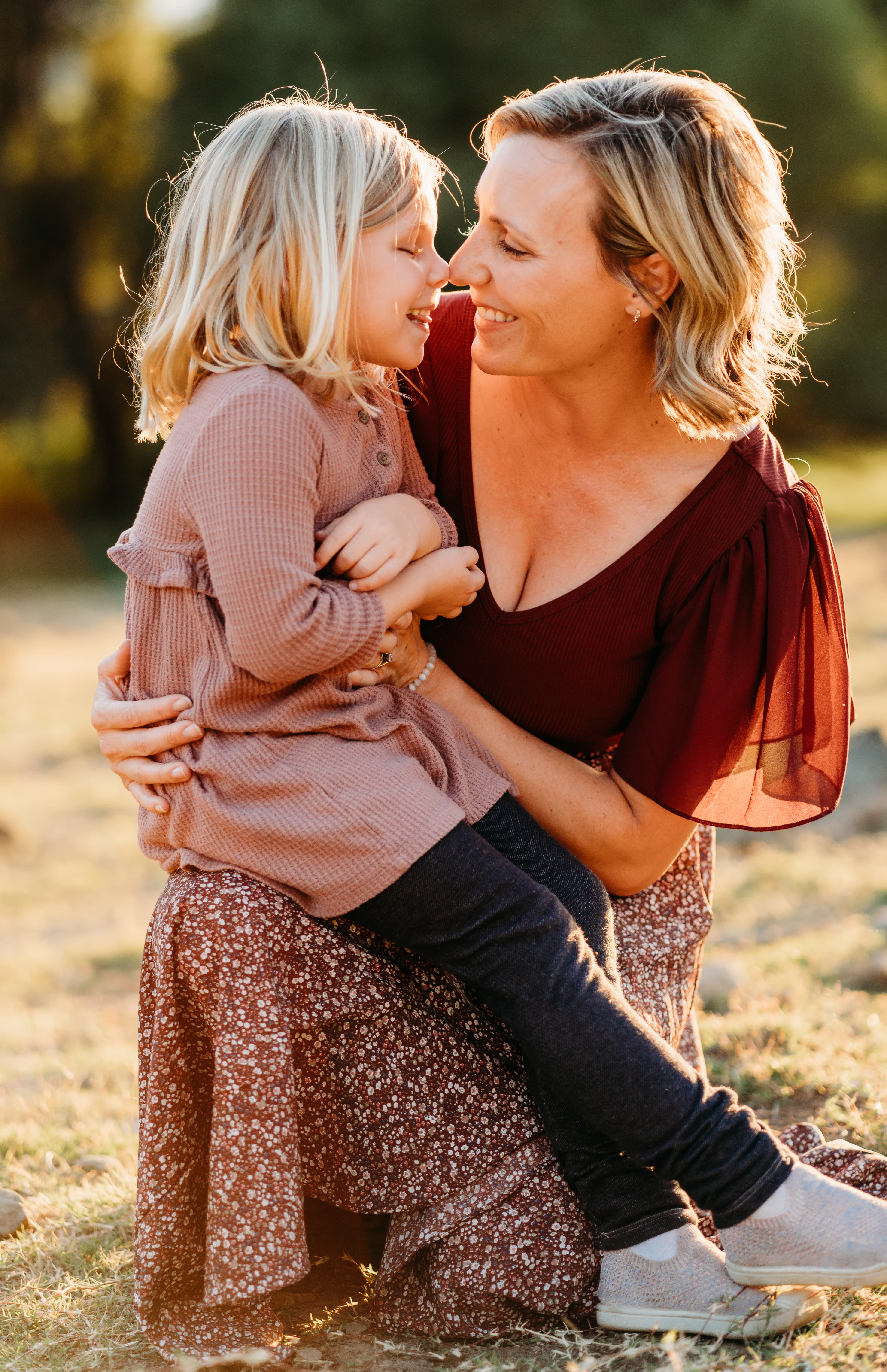 Mother in maroon top and her young blonde daughter touch noses, laughing