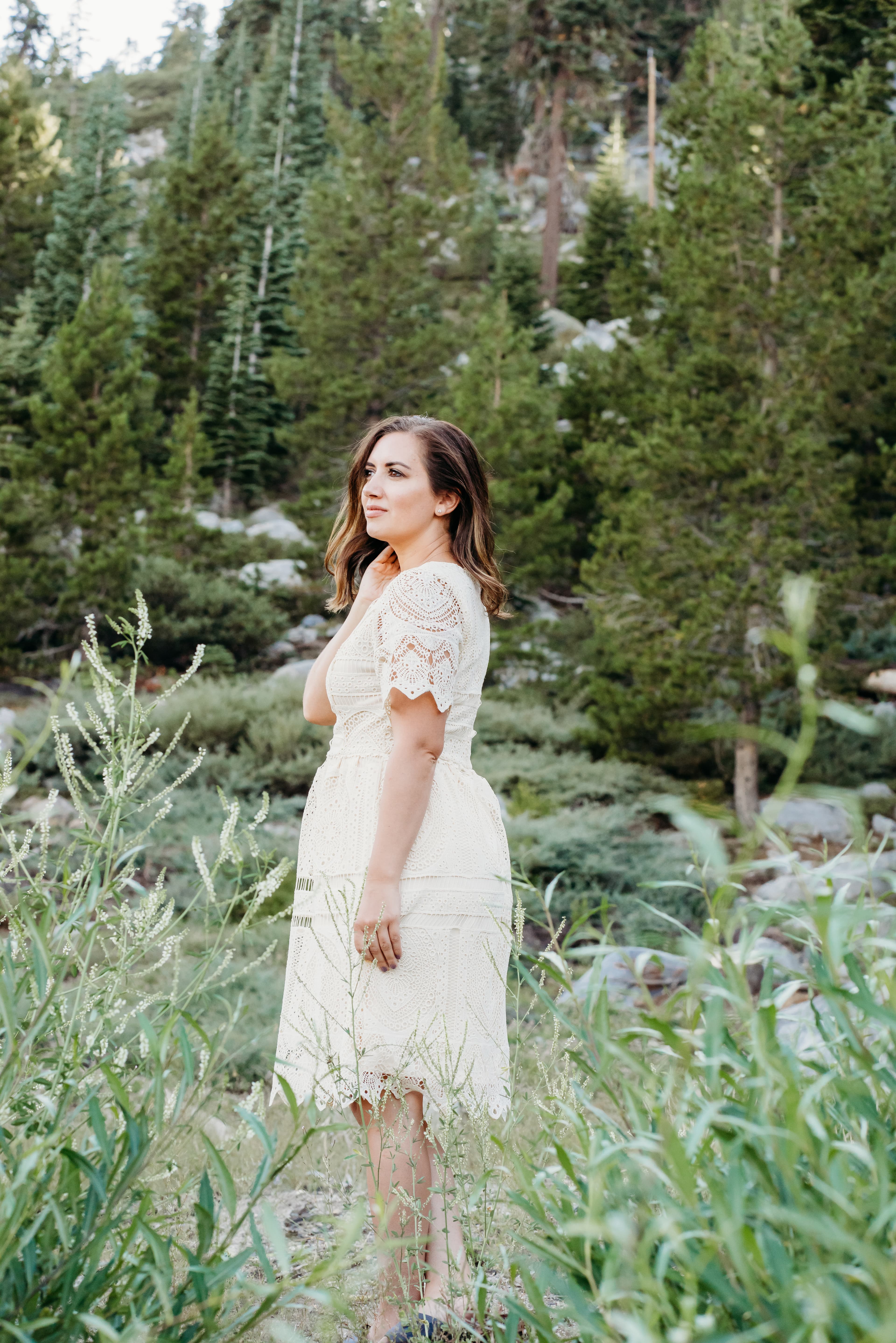 Woman in a white lace dress stands among pine trees, touching her hair