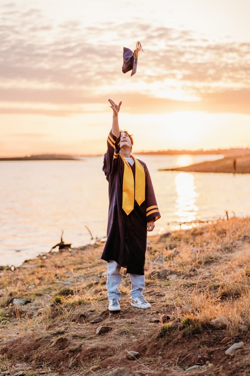 Graduate in cap and gown tosses his cap into the air beside a sunset lake