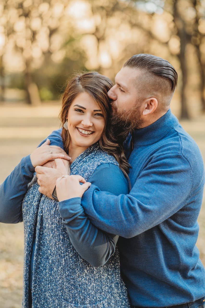 Man in a blue sweater kisses his smiling partner's head, both embracing outdoors