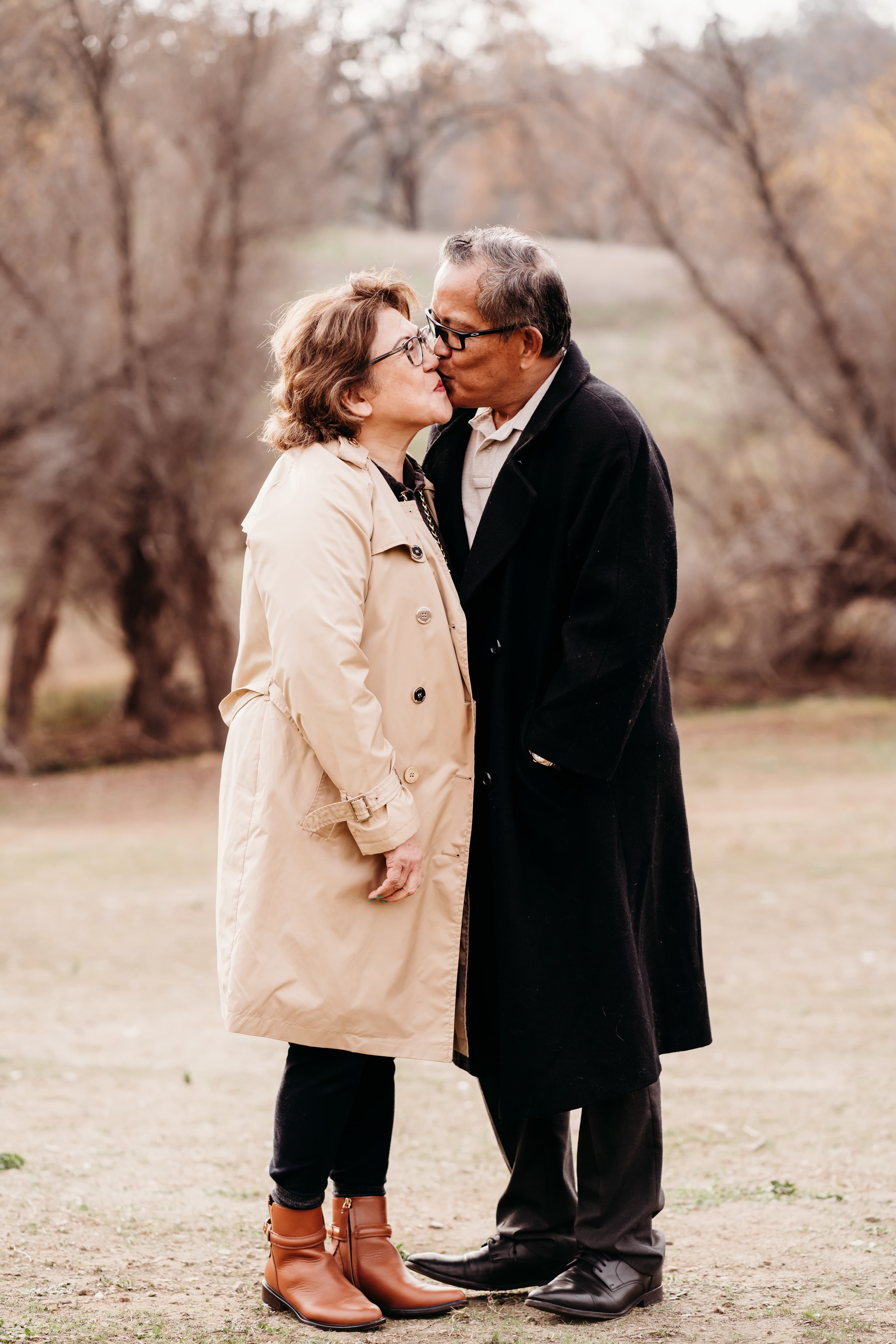 Older couple shares a kiss outdoors in coats among bare autumn trees