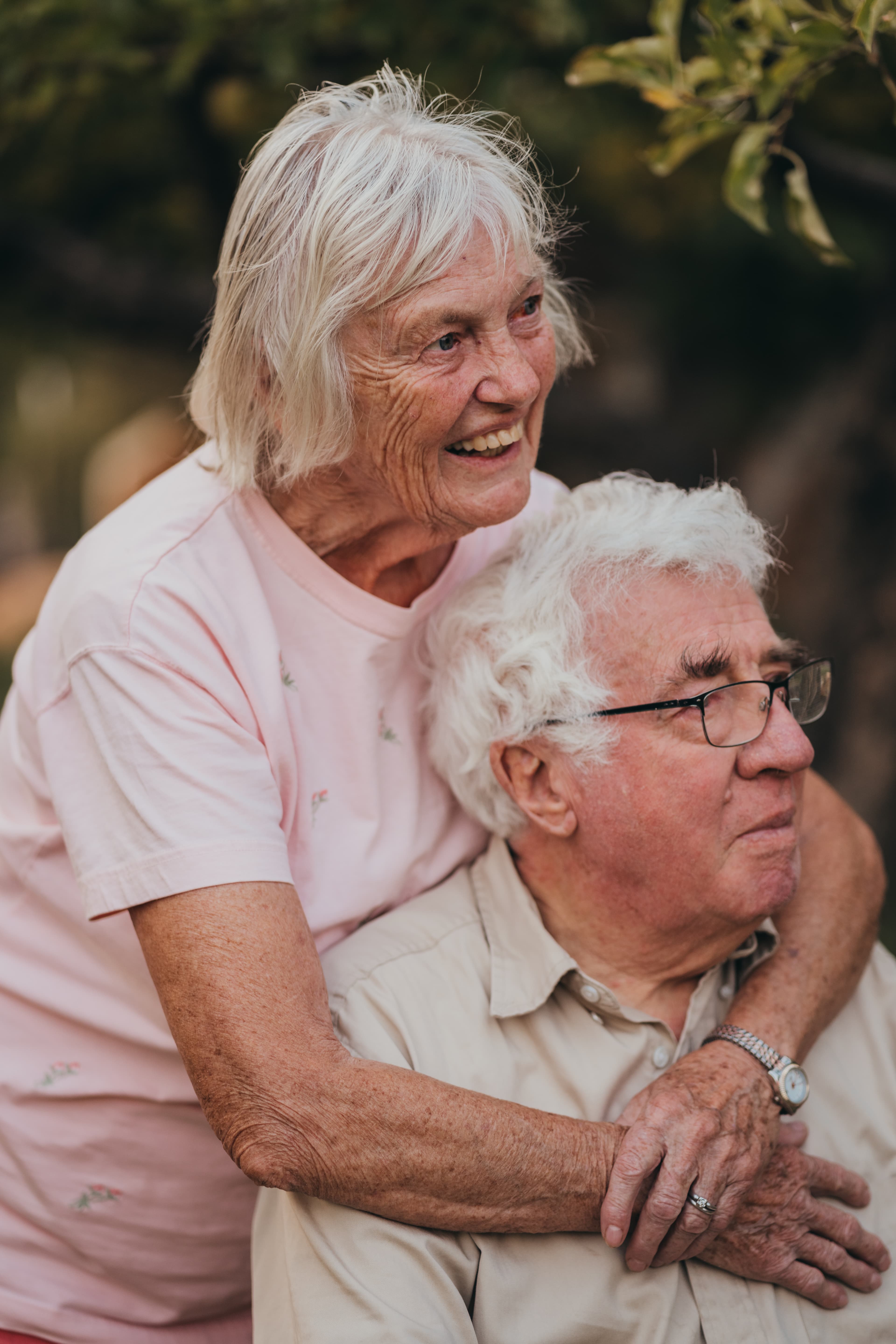 Elderly couple embrace outdoors, she laughing in a pink shirt