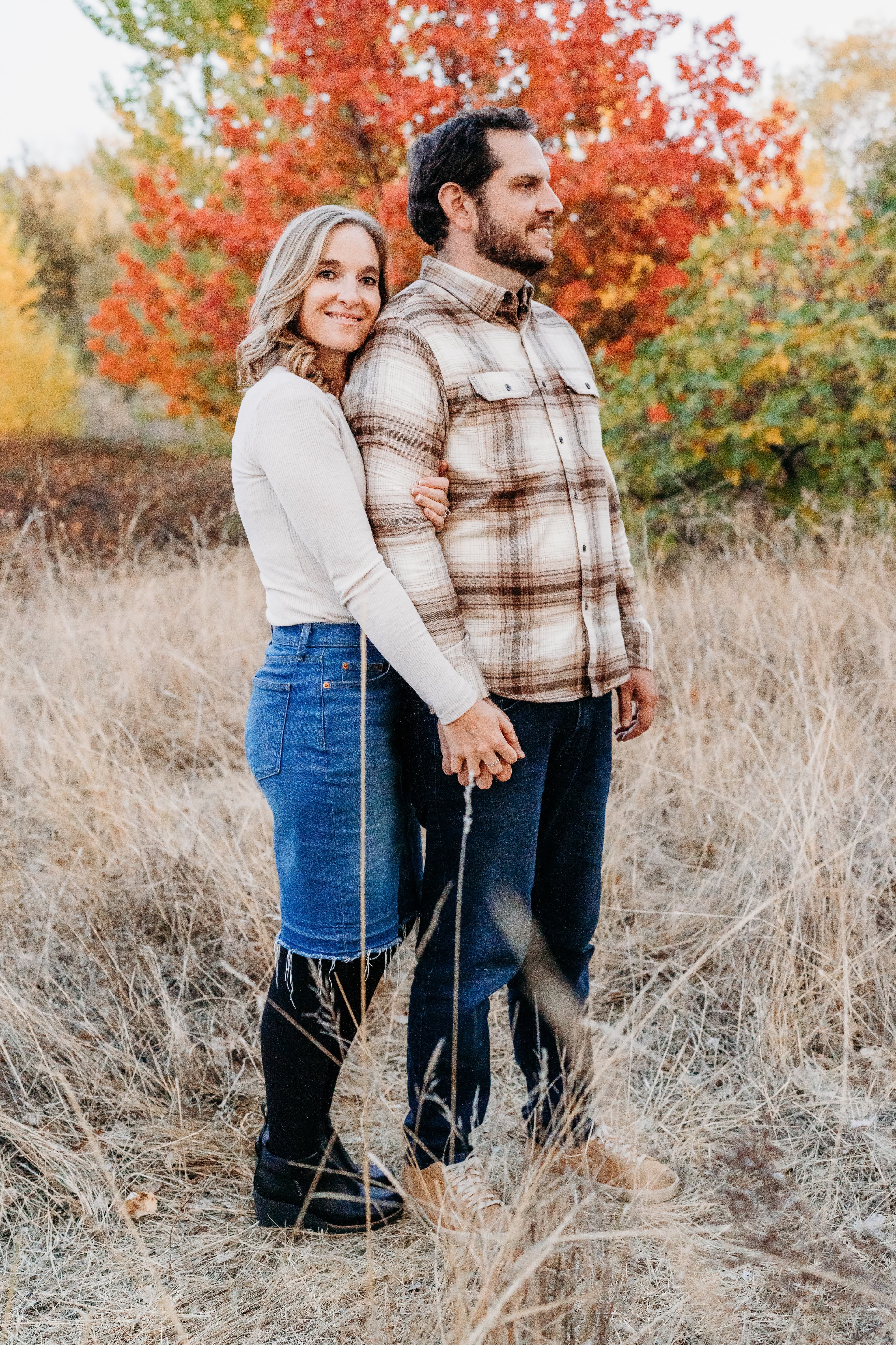 Woman embraces a man from behind in a golden grass field with vibrant red autumn foliage behind them