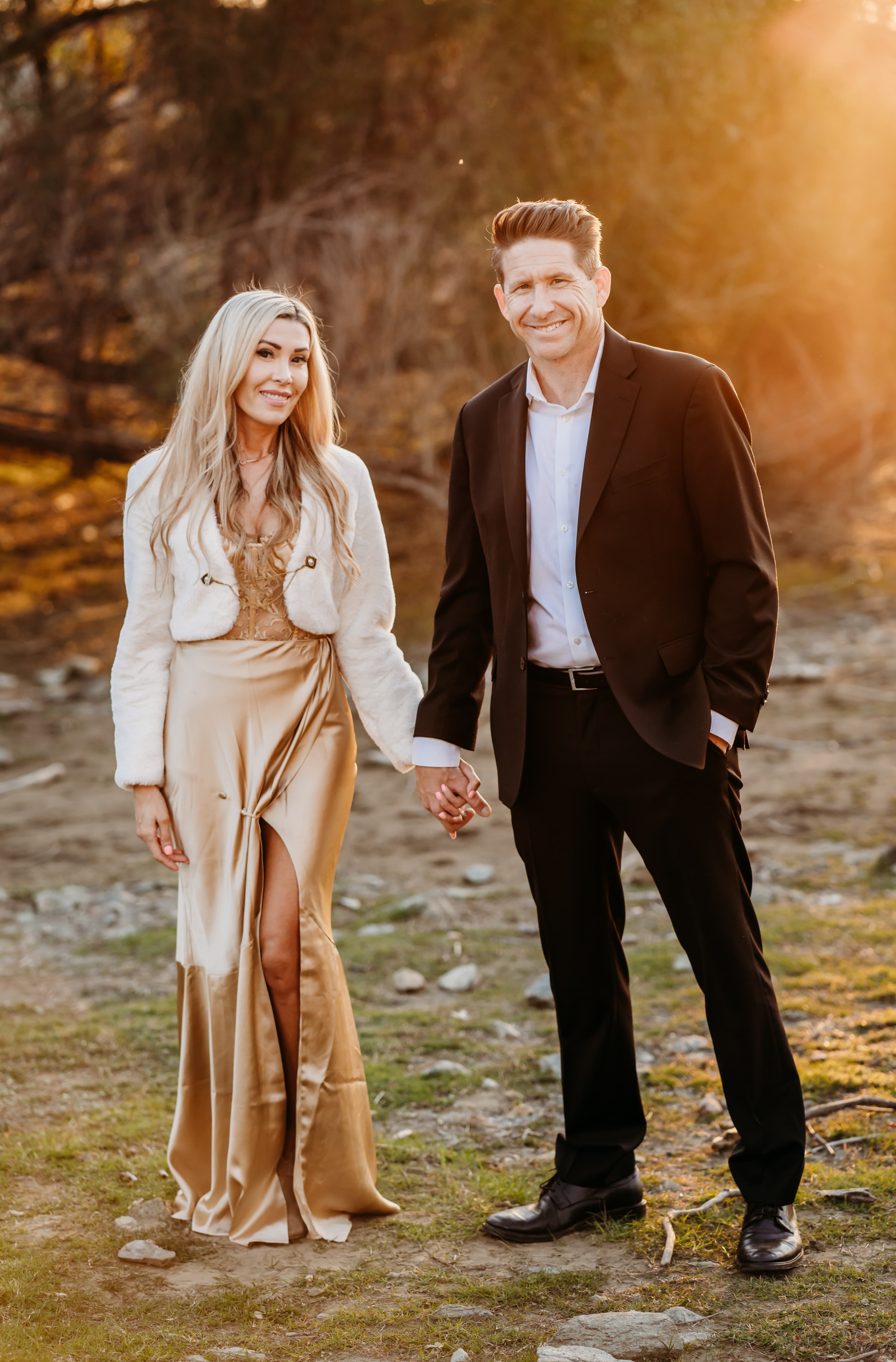 Couple holds hands and smiles, glowing in warm golden-hour backlight