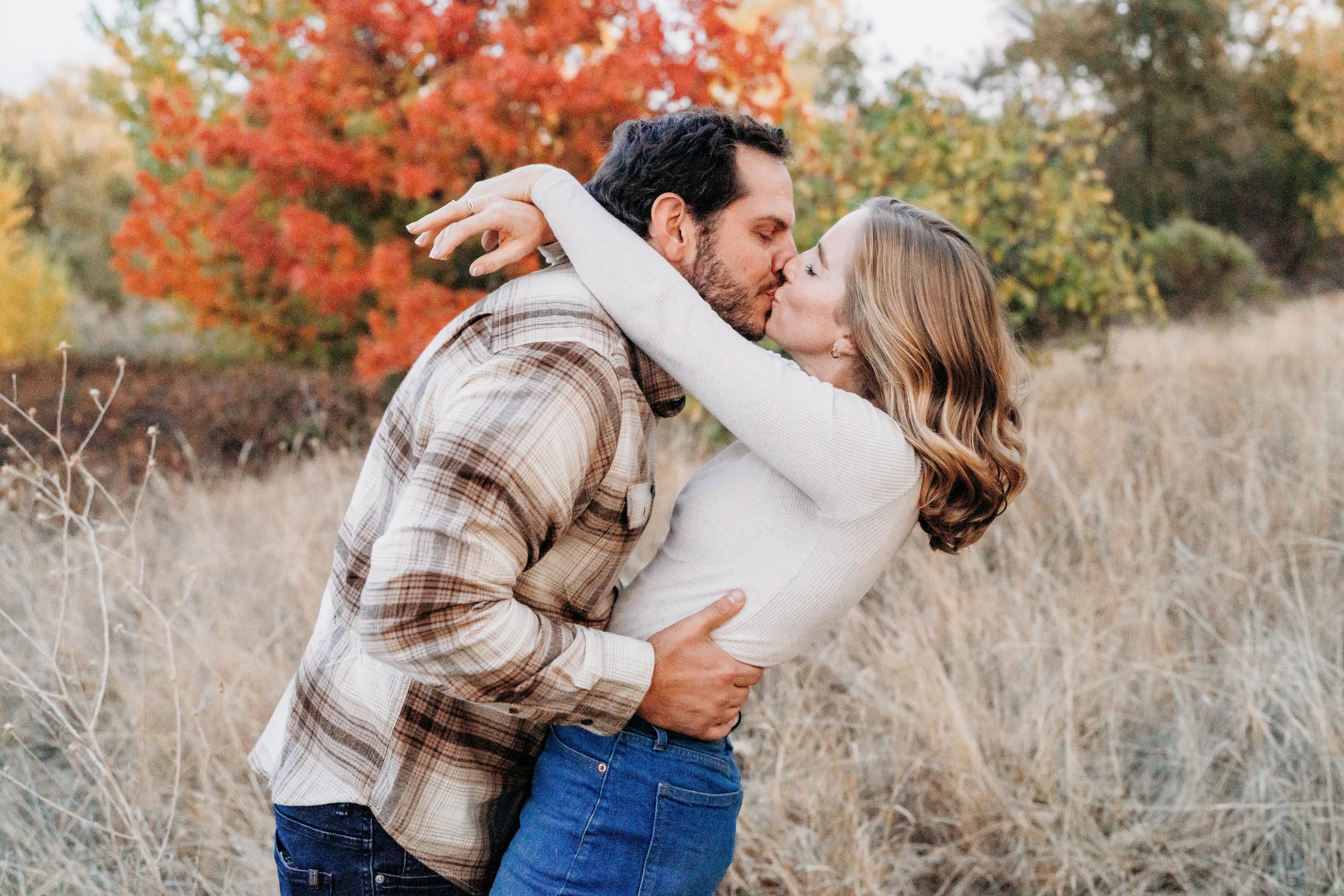 Couple shares a kiss in a dry grass field with autumn trees in the background