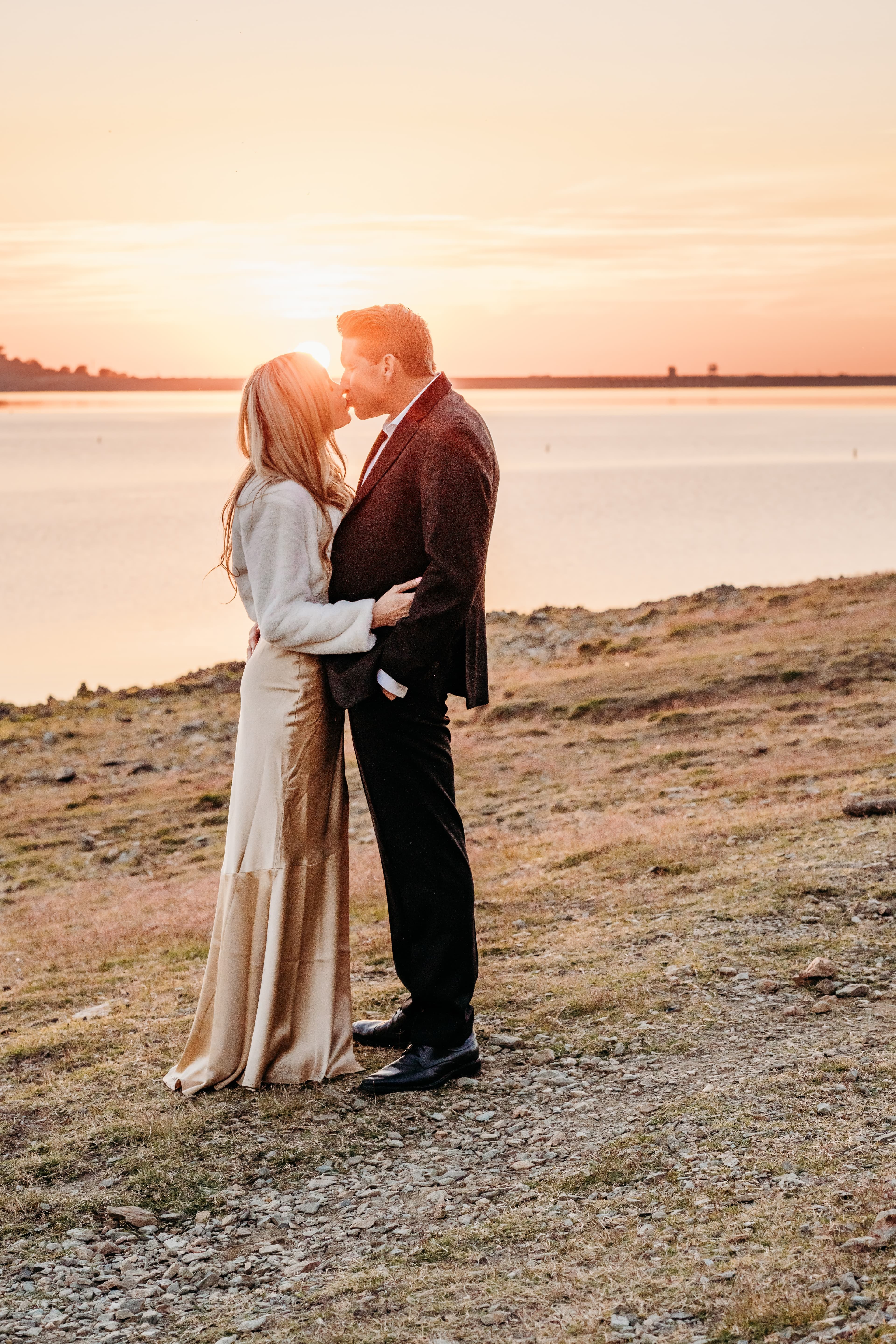 Couple kisses at the edge of a lake, silhouetted against a warm sunset