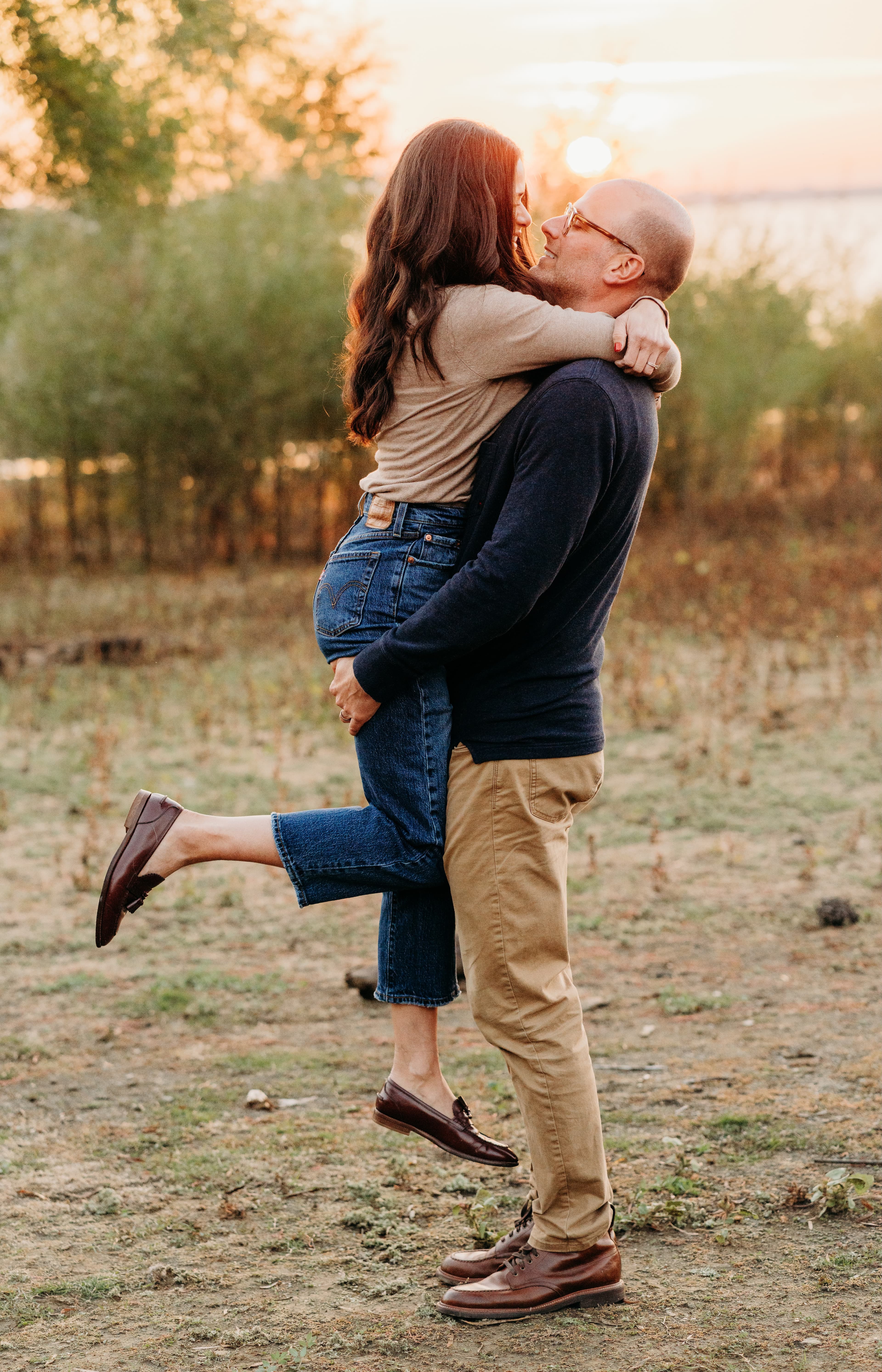 Man joyfully lifts his partner off the ground by the lake at sunset