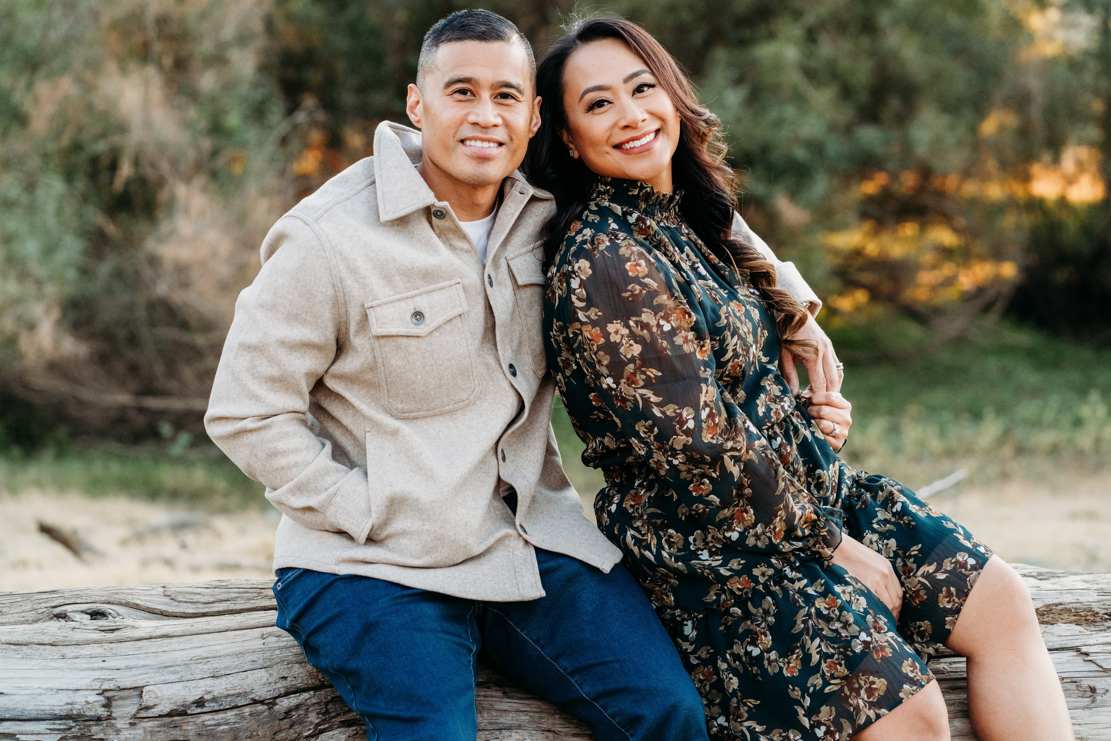 Couple sits close together on a fallen log in the woods, smiling at the camera