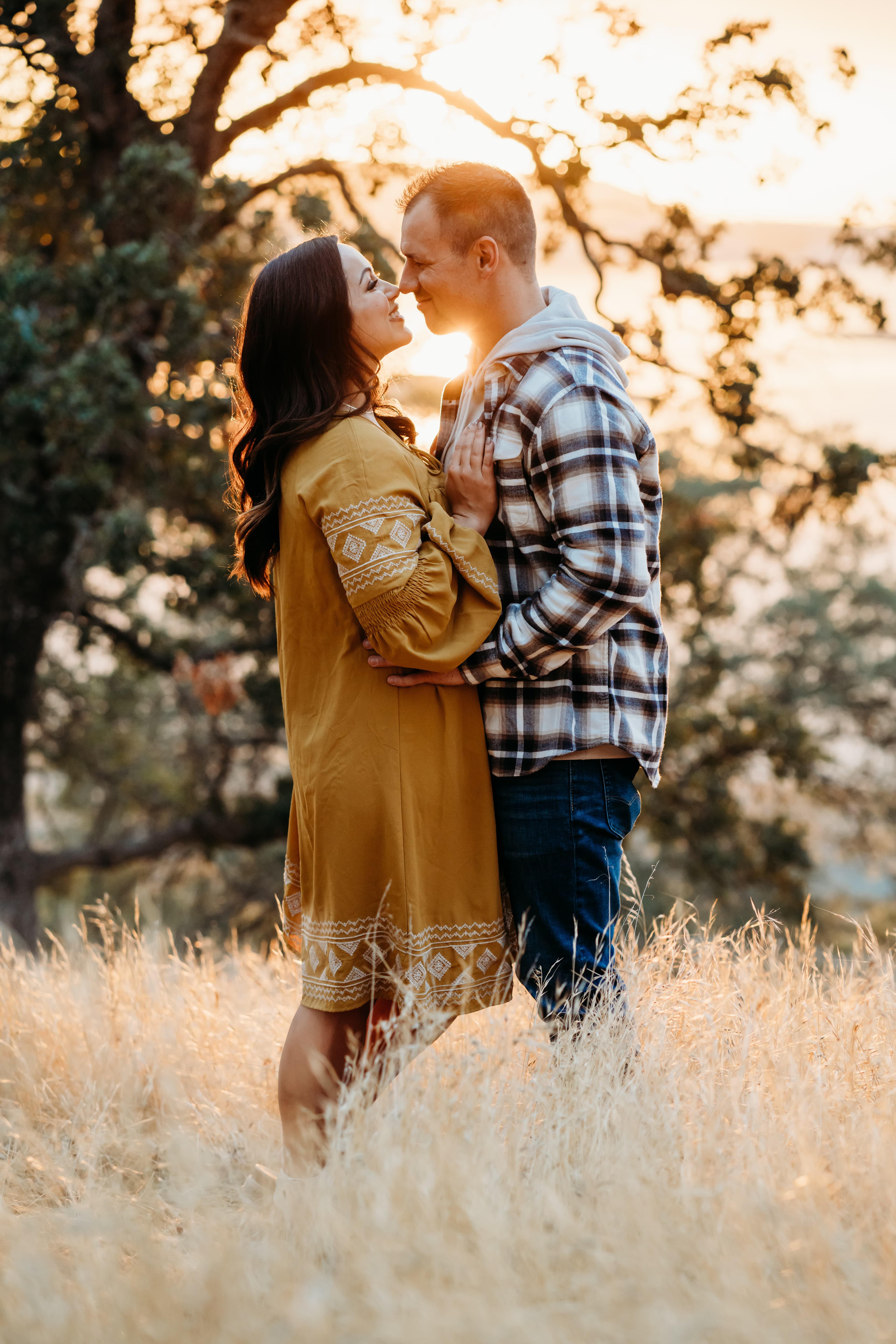 Couple nearly kiss nose to nose, she in mustard, he in flannel, sunset tree