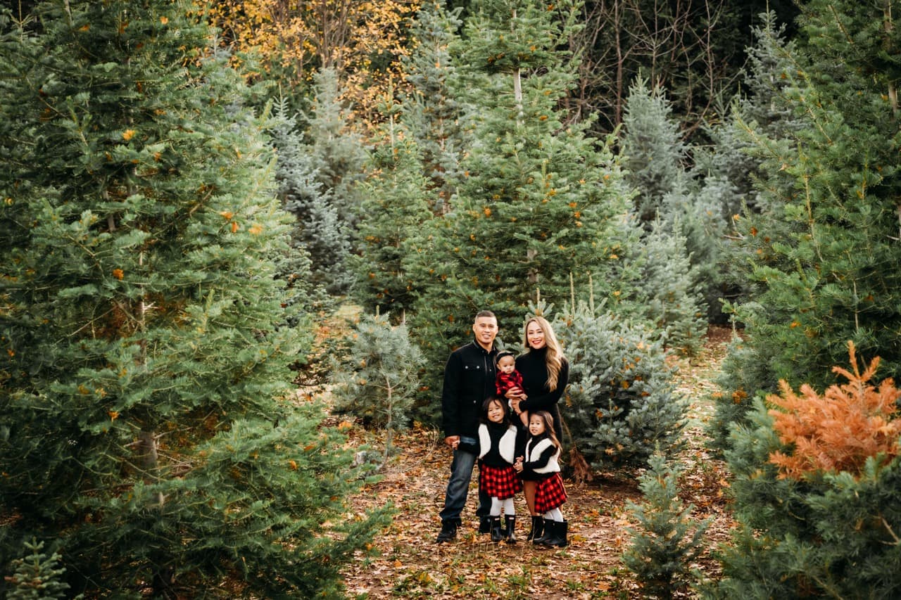 Family of five in black and red plaid stand together in a Christmas tree farm