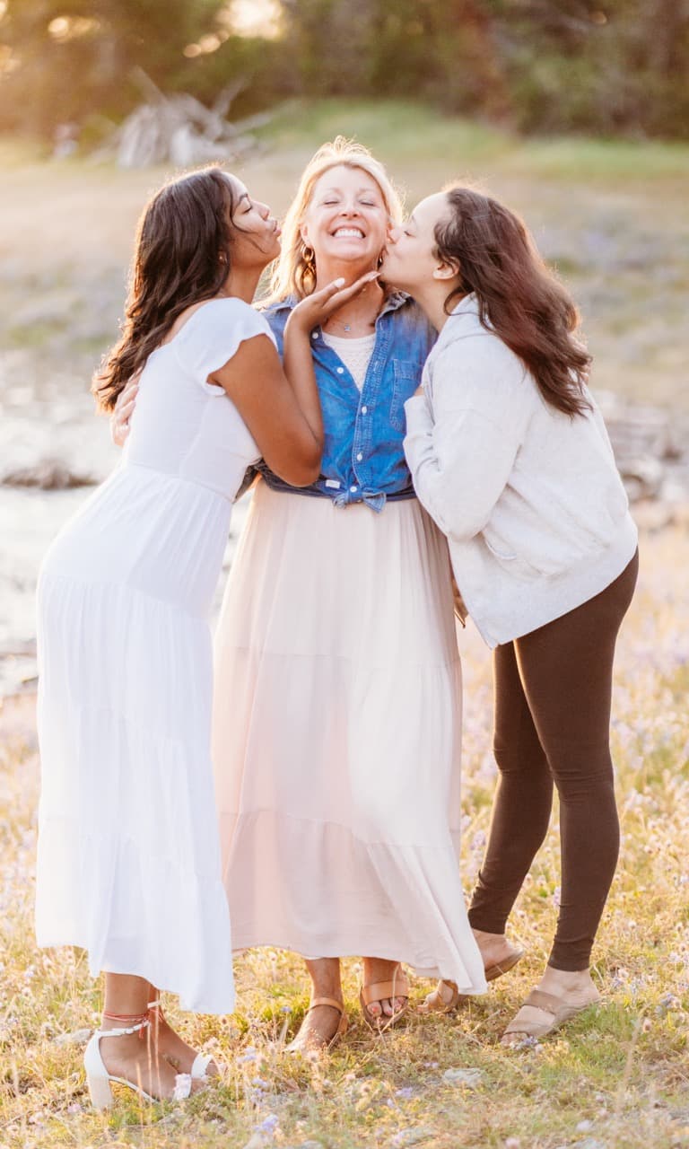 Two young women kissing an older woman's cheeks in a wildflower field