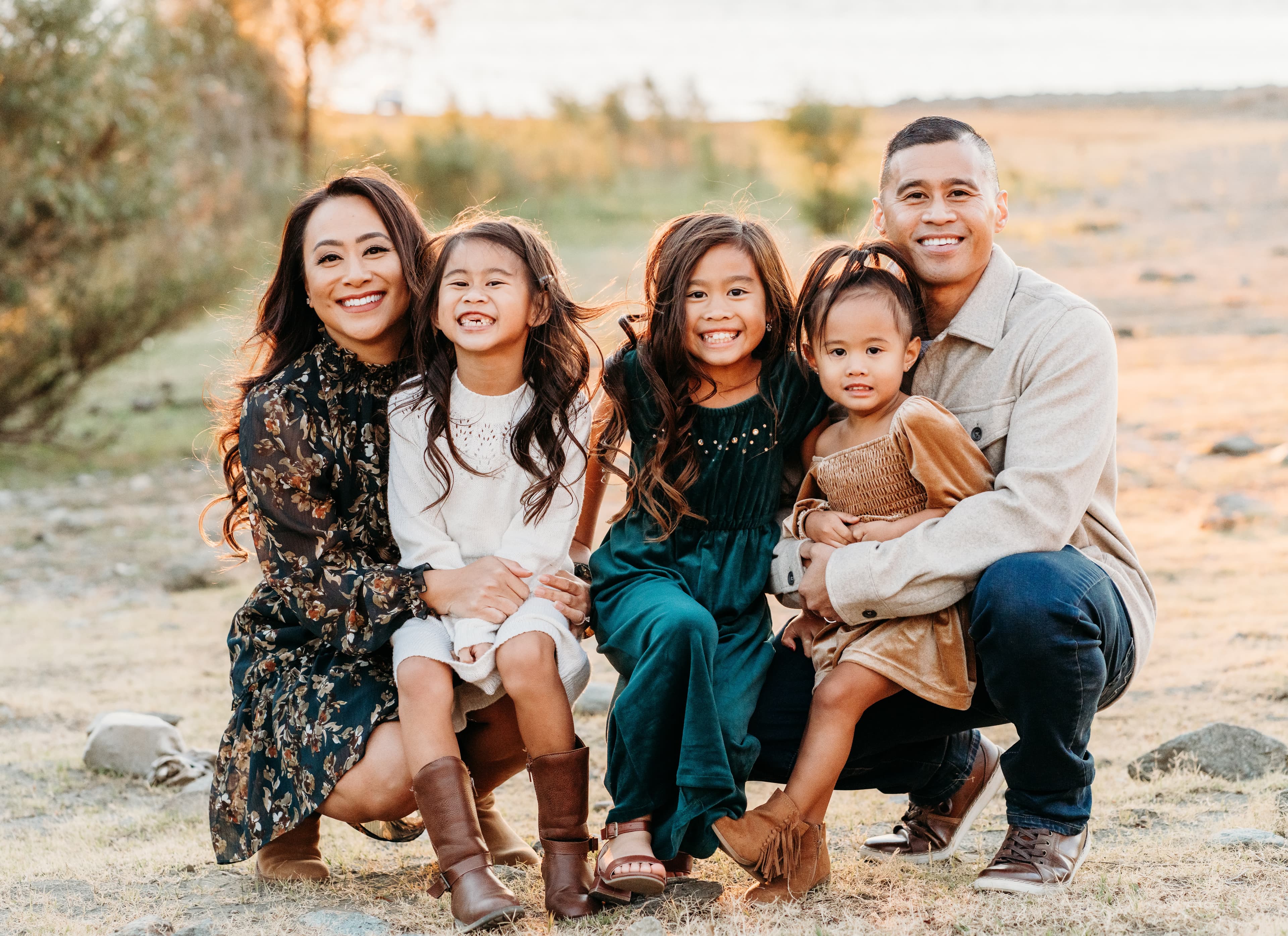 Family of five crouches together for a portrait in a golden field near the water at sunset