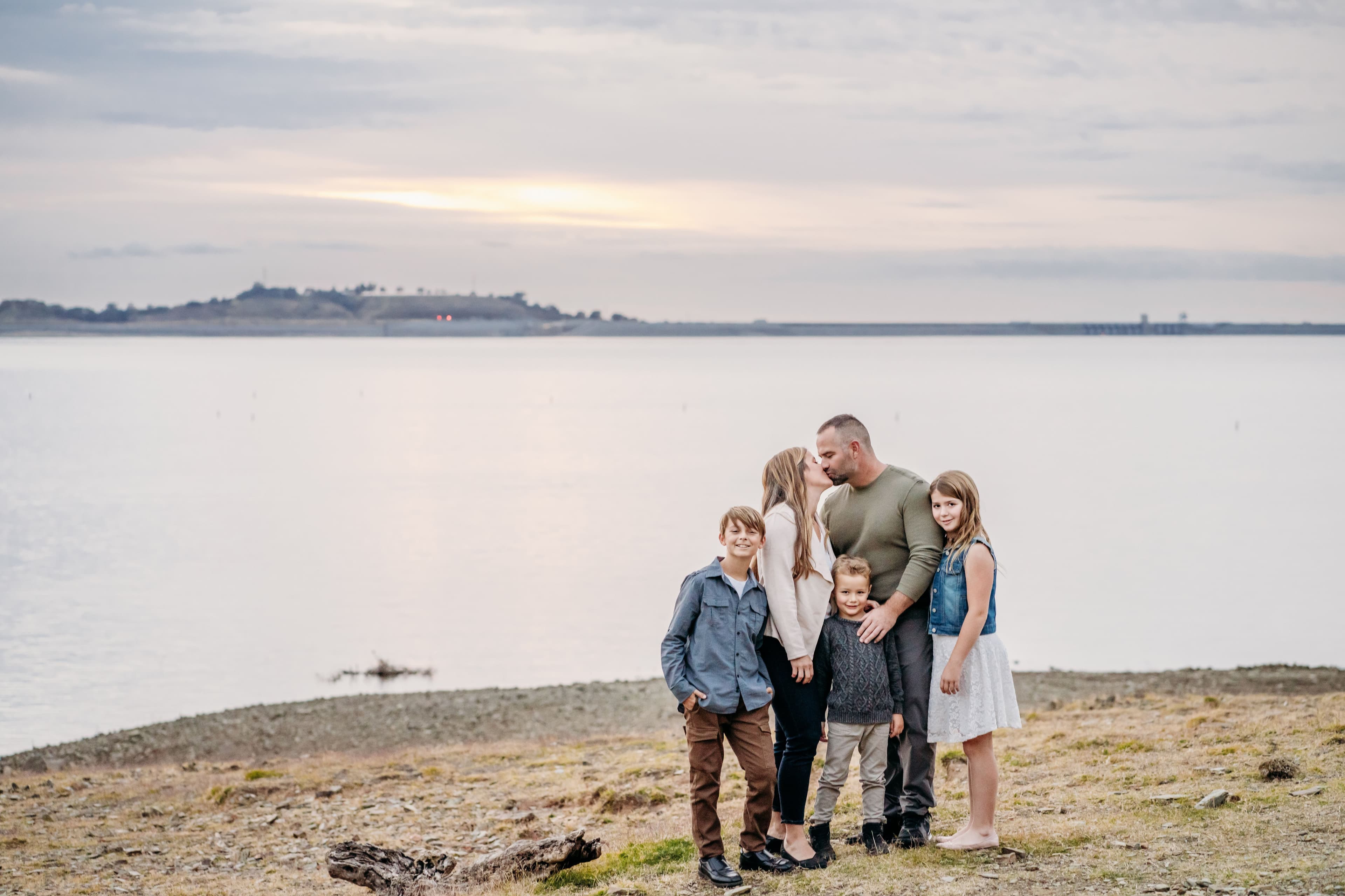 Family of five stands on a lakeside bluff at dusk, parents sharing a kiss