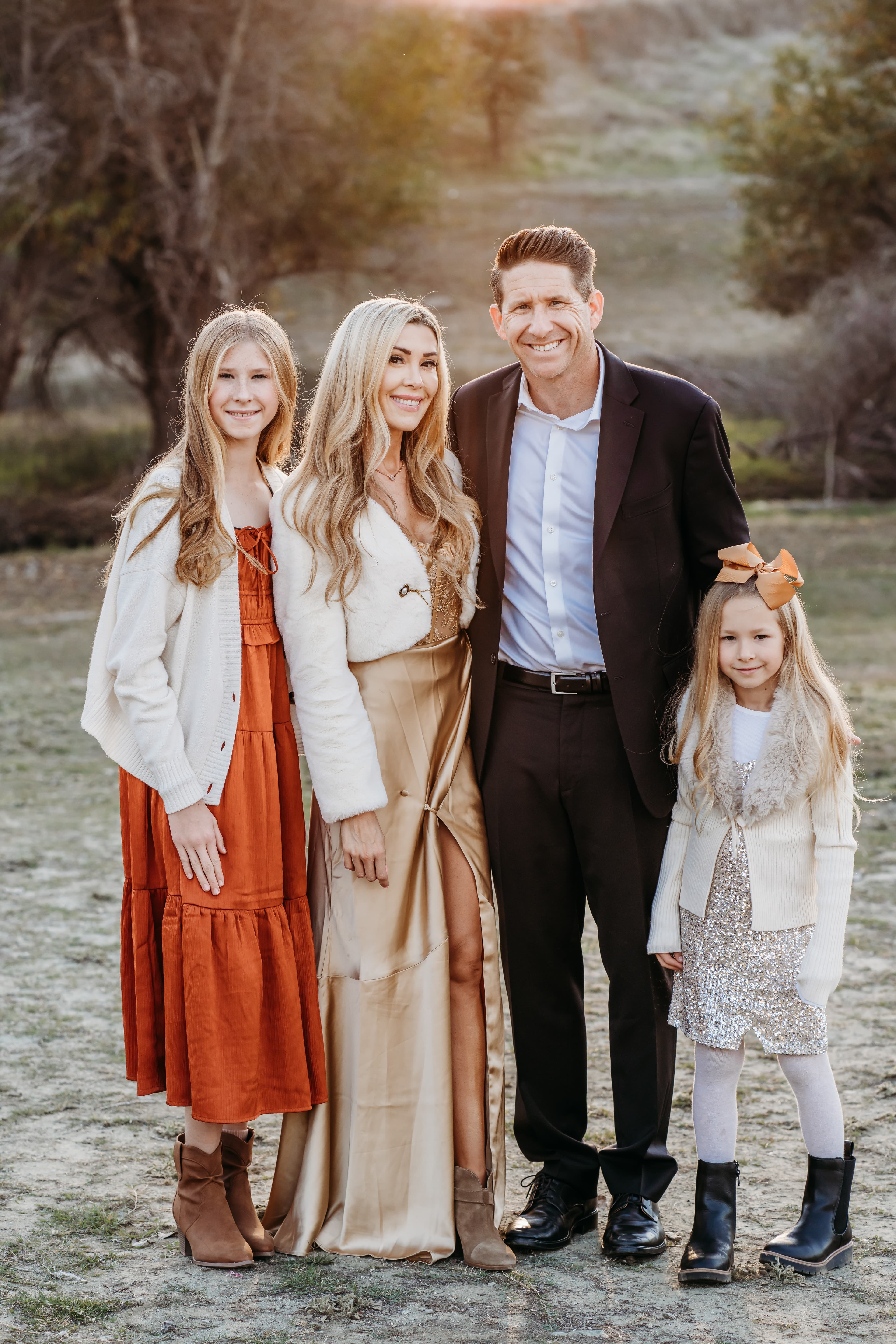 Family of four in warm-toned formal attire stands together in a dry grass field