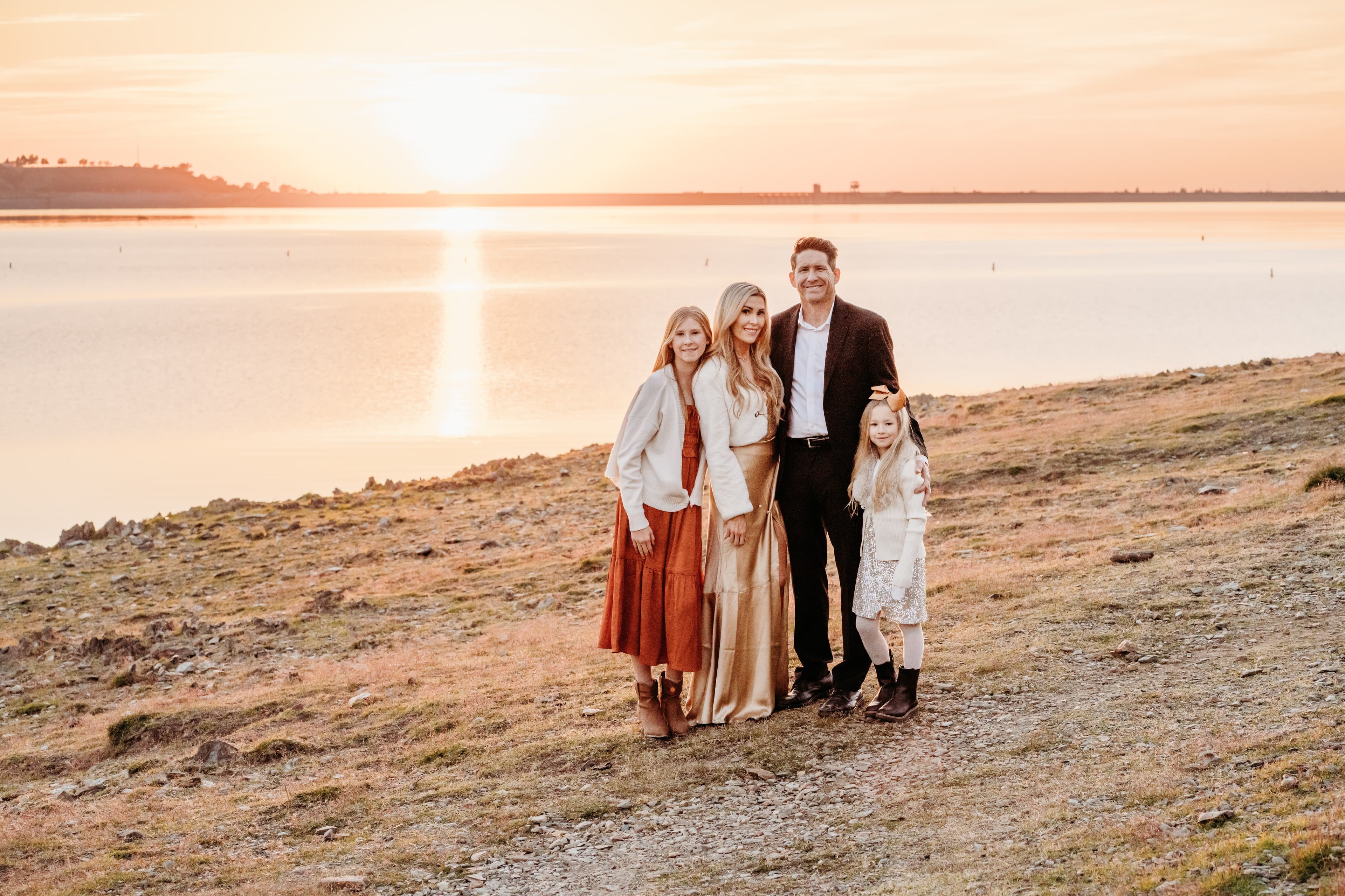 Family of four stands together on a rocky hillside overlooking a lake at golden-hour sunset