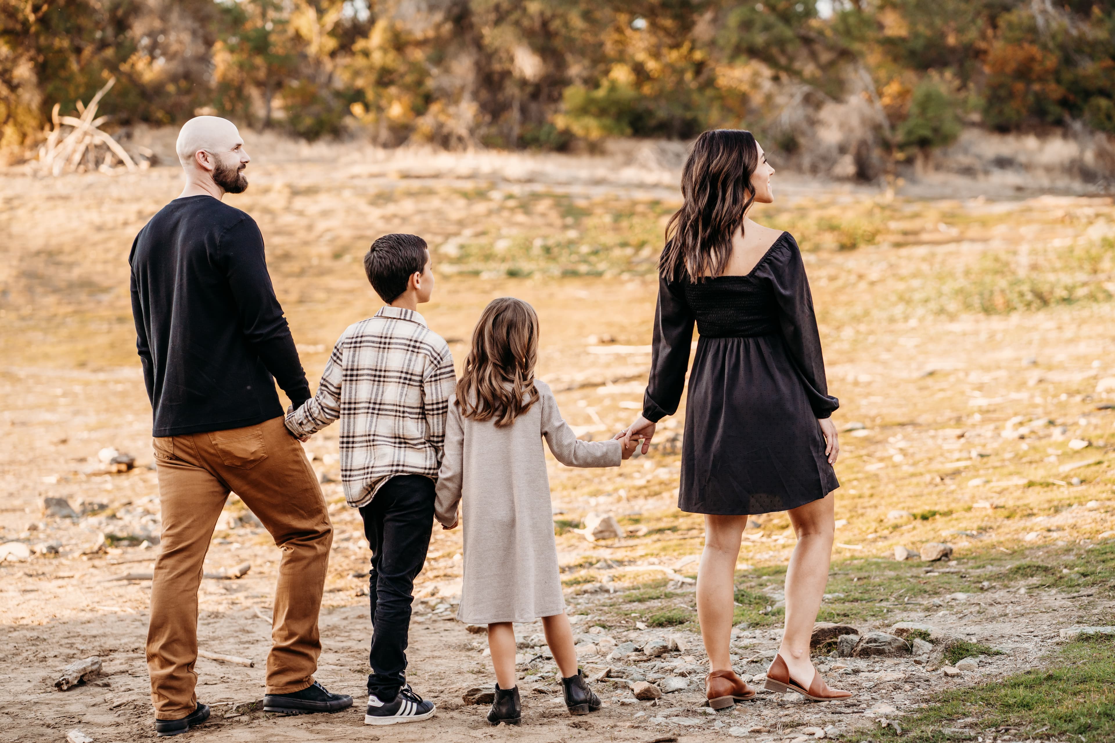 Family of four walks hand in hand away from the camera through a golden autumn field, glancing back and smiling