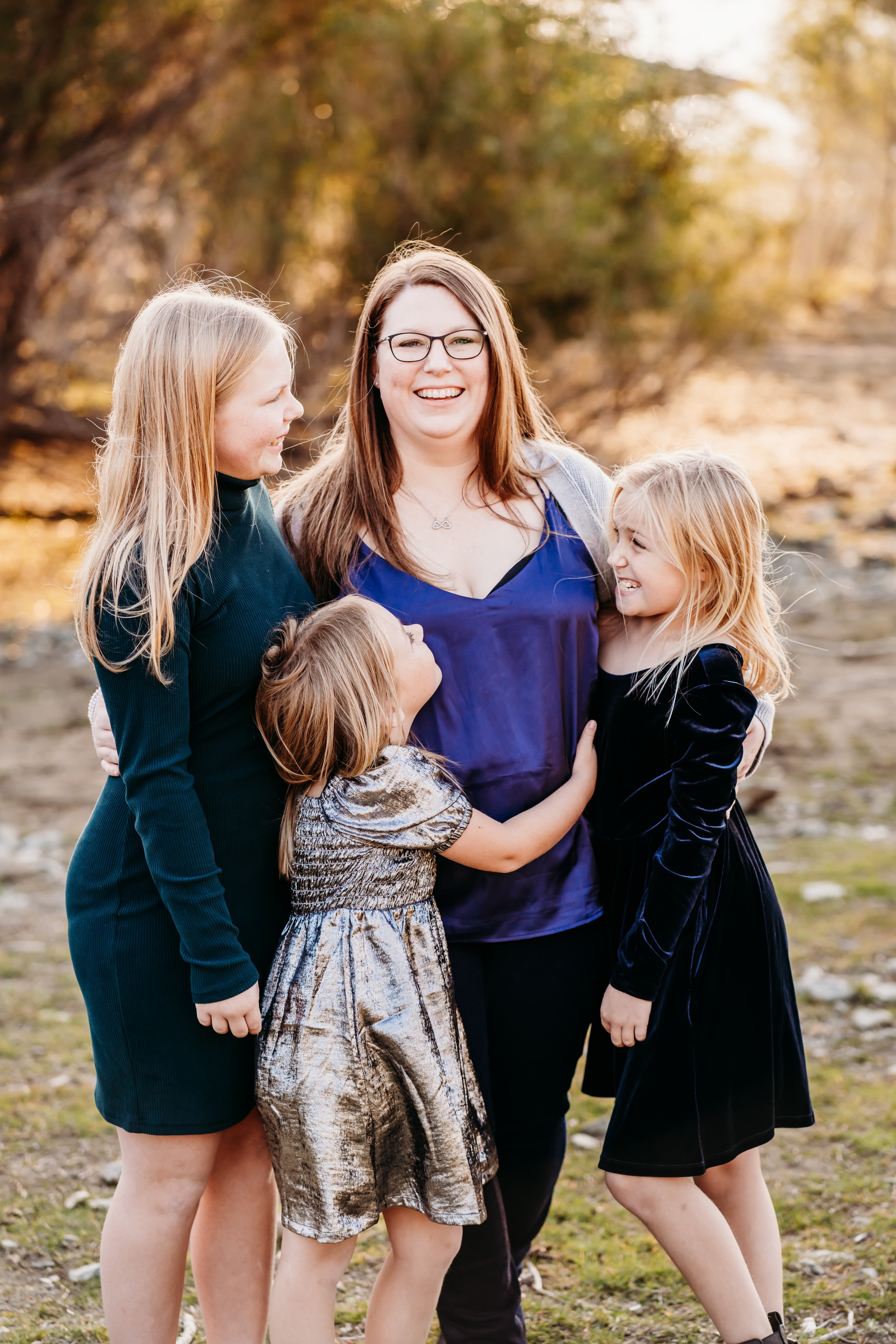 Mother laughs joyfully while hugged by her three daughters outdoors at sunset
