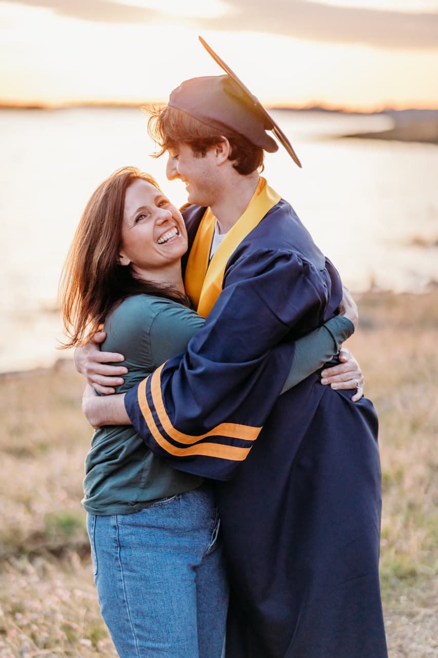 Mother laughing while hugging her son in cap and gown by a lake