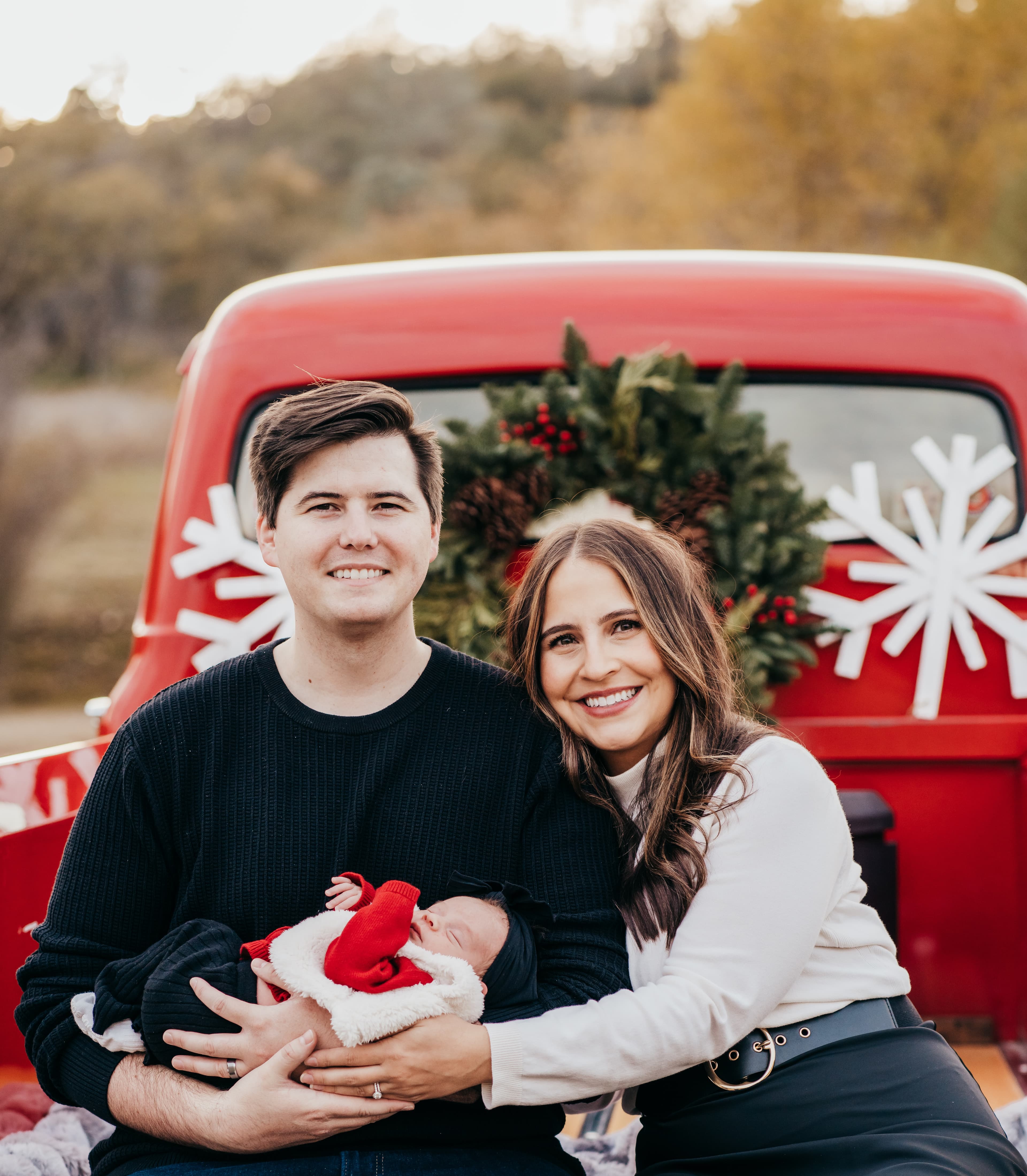 New parents hold their sleeping newborn while sitting on a vintage red truck decorated for Christmas