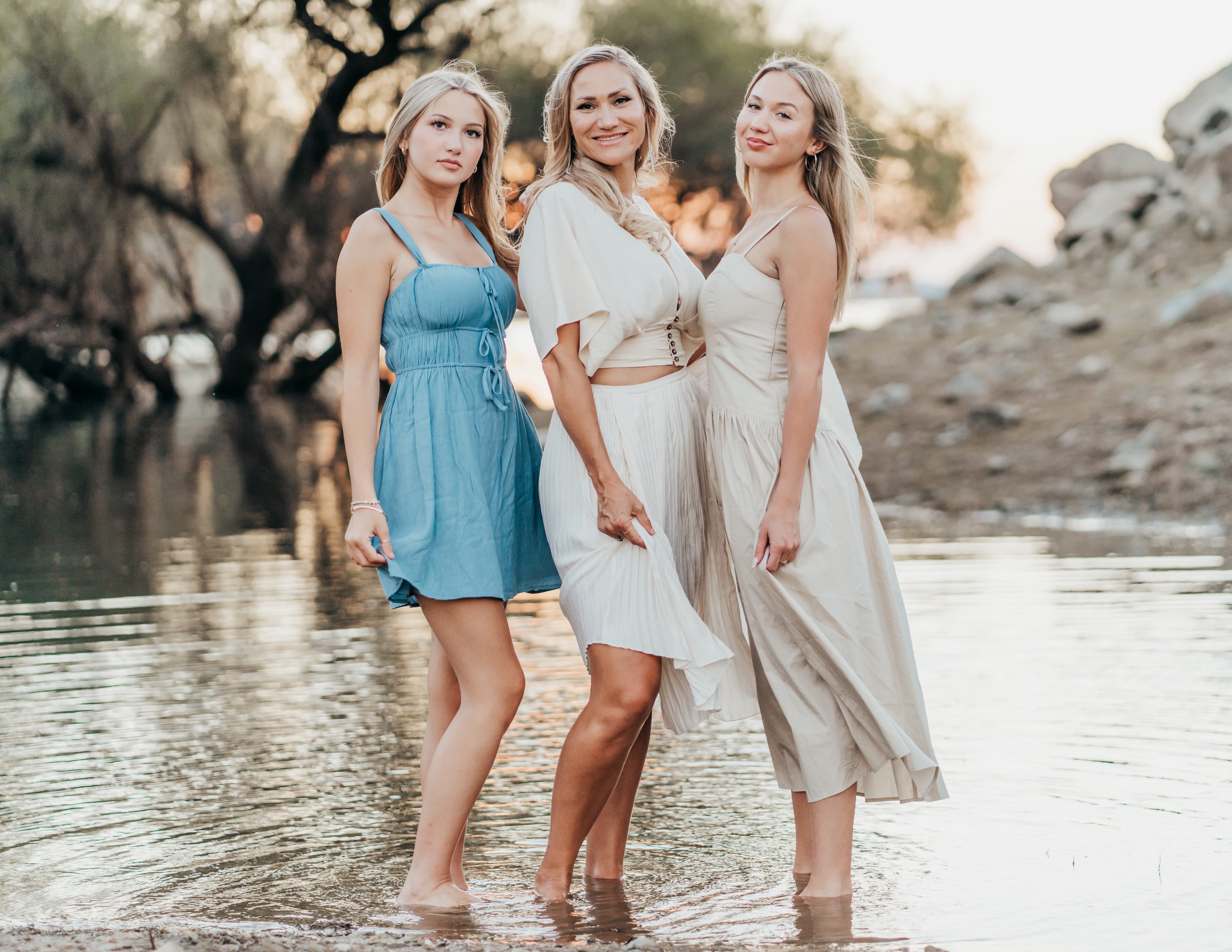Three generations of women stand ankle-deep in a river at golden hour, smiling at the camera