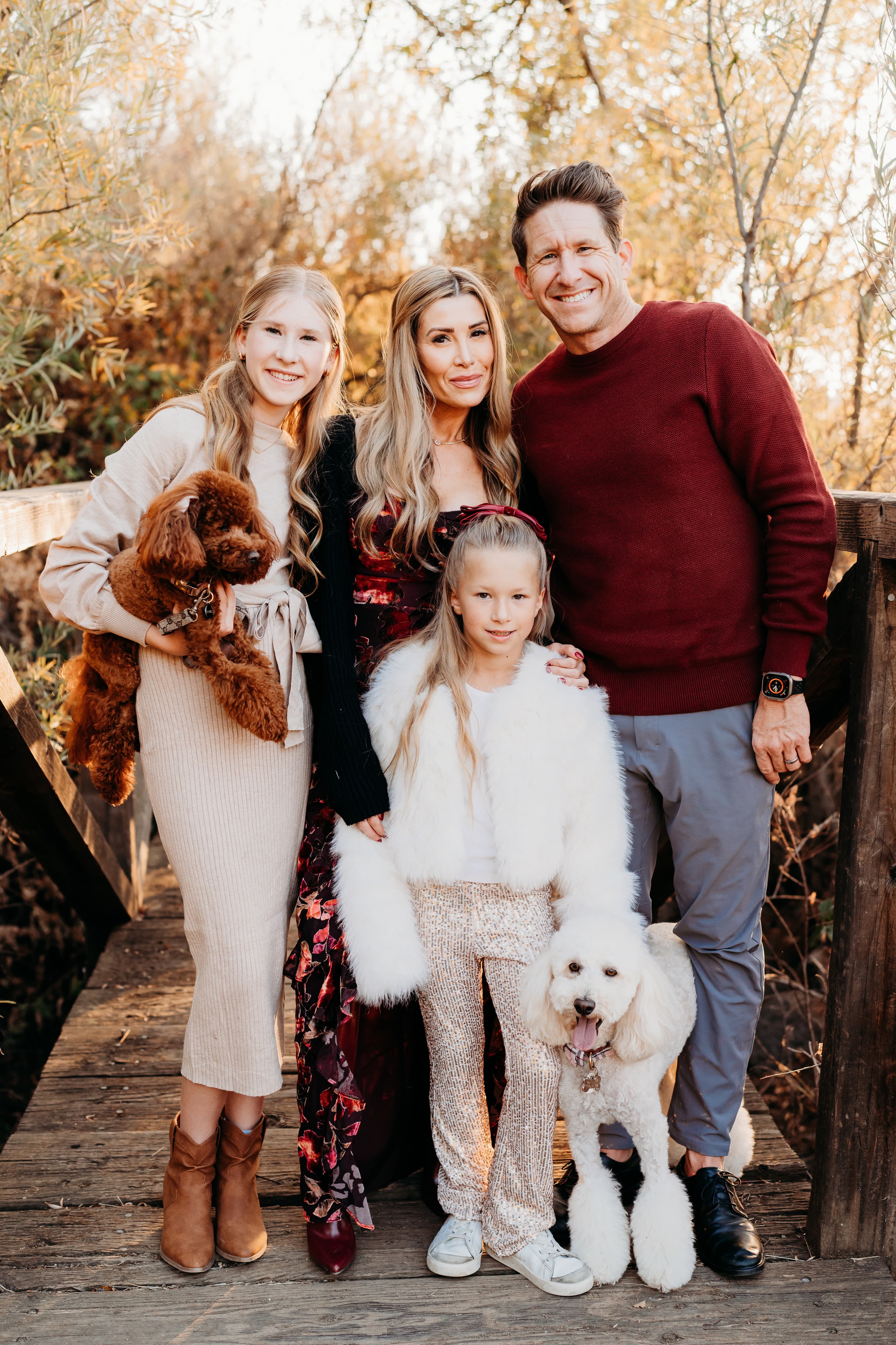 Family of four poses on a wooden bridge with their two poodles amid autumn leaves