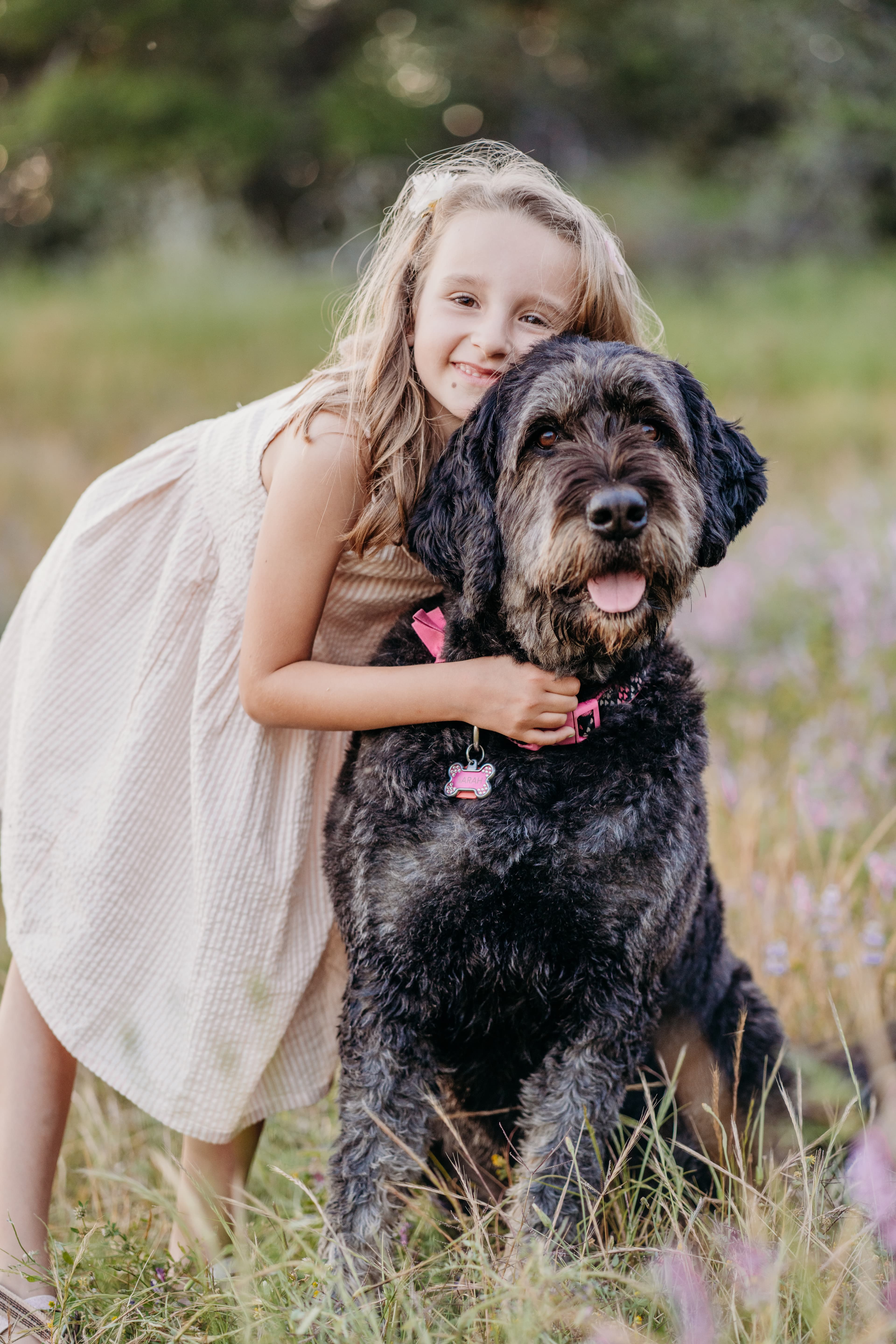 Smiling young girl rests her cheek on her dog's head while standing in a wildflower field