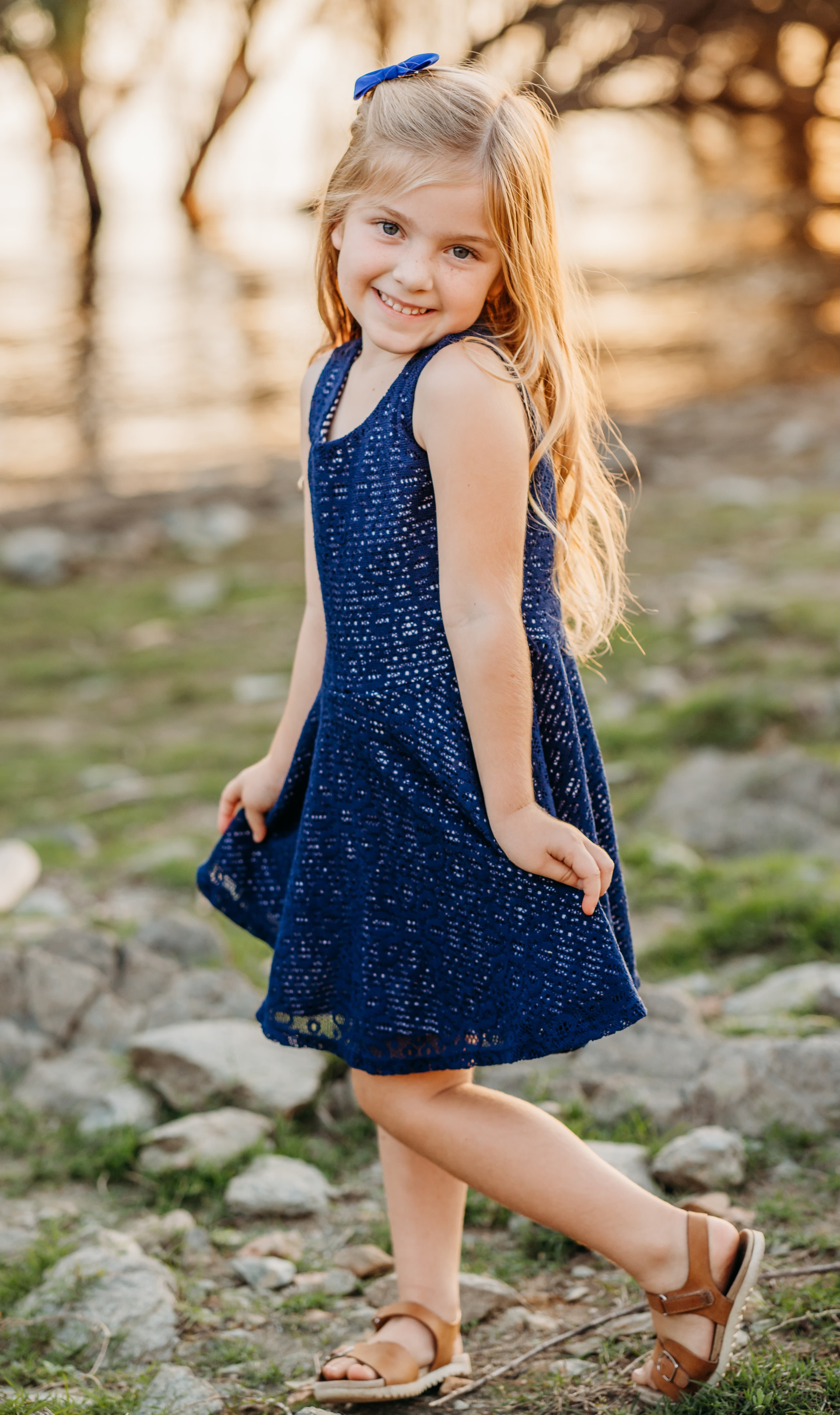 Young girl in a navy dress twirls and smiles on a rocky lakeshore at sunset