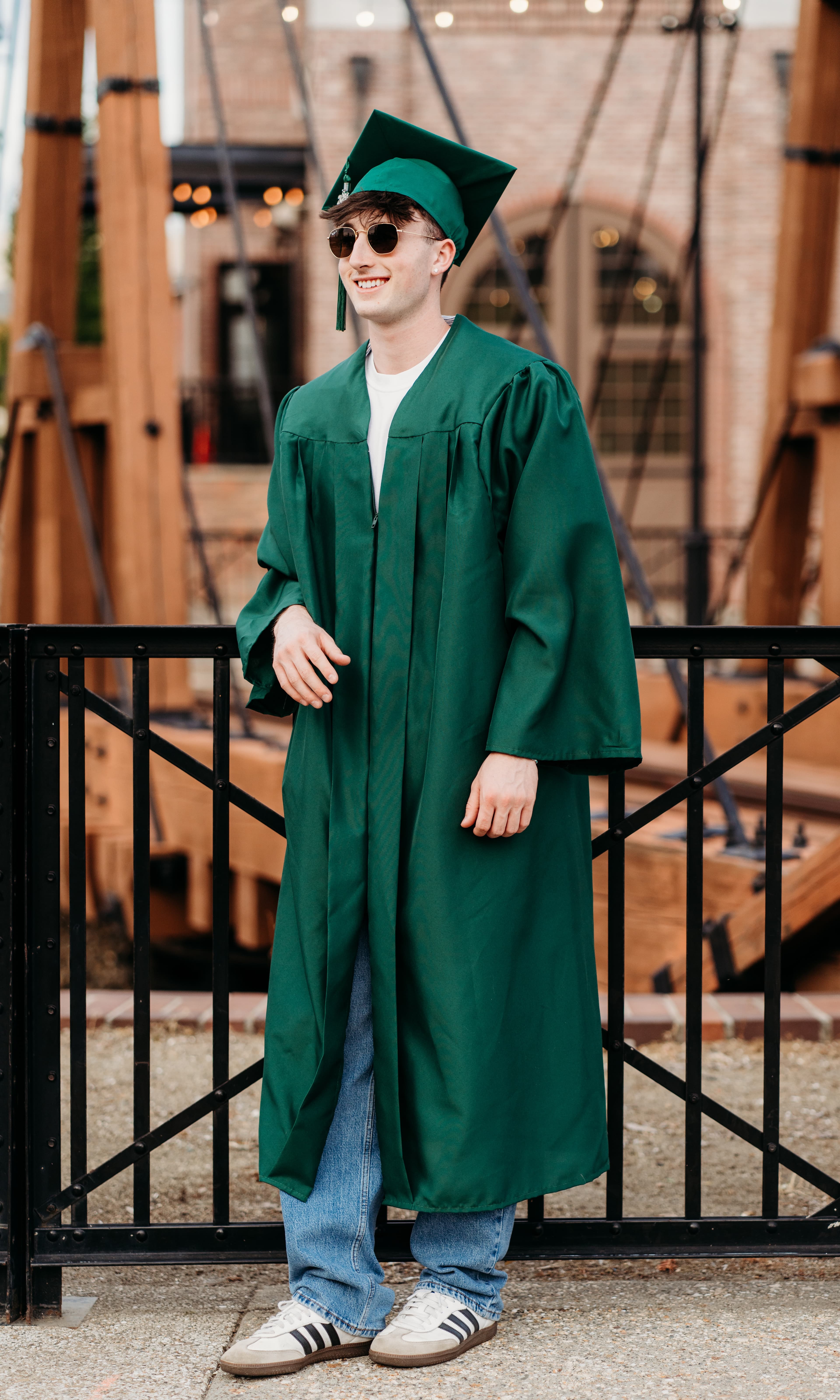 Graduate in a green cap and gown wearing sunglasses leans against a railing, smiling