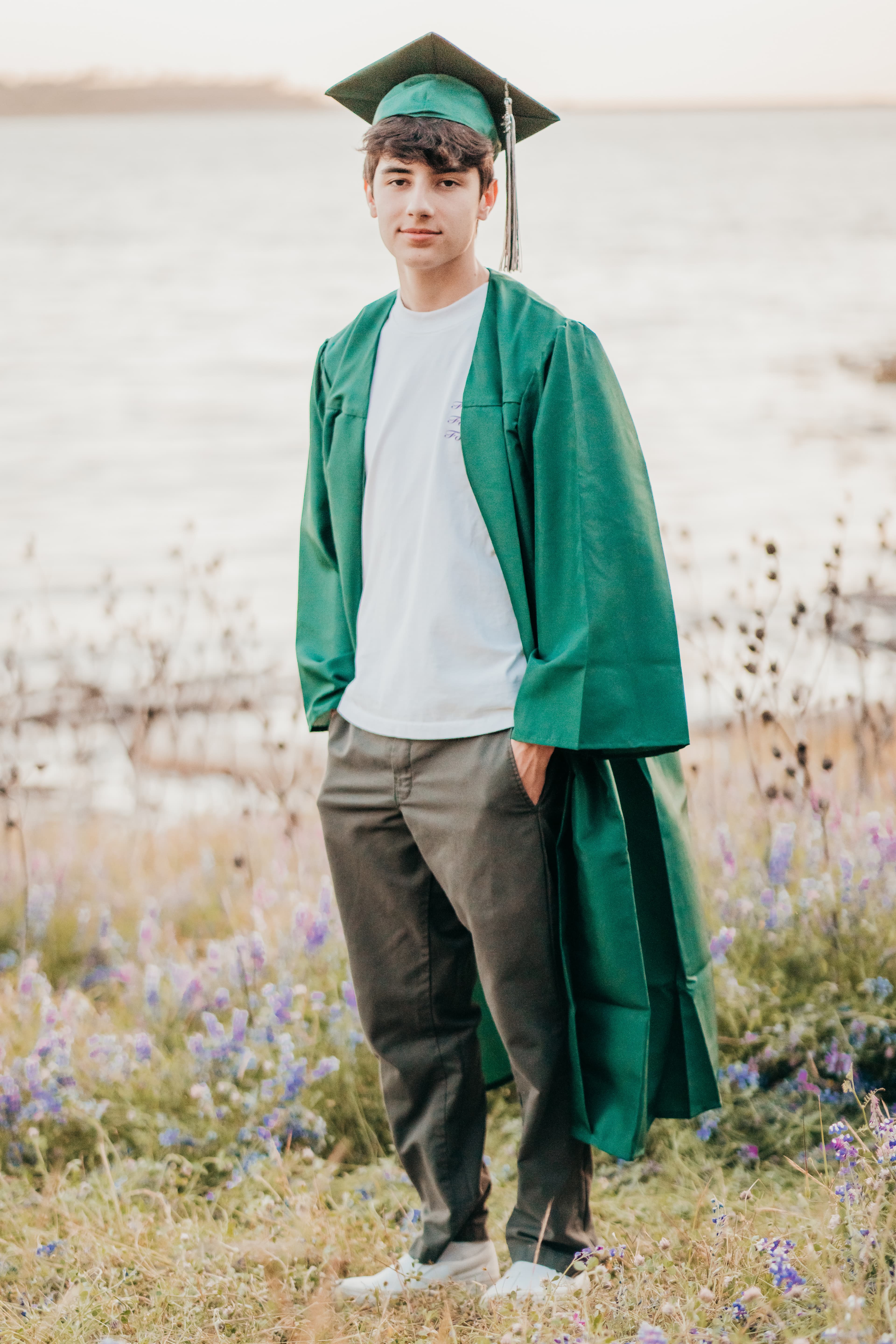 Teen boy in a green cap and gown stands in a wildflower field by a lake