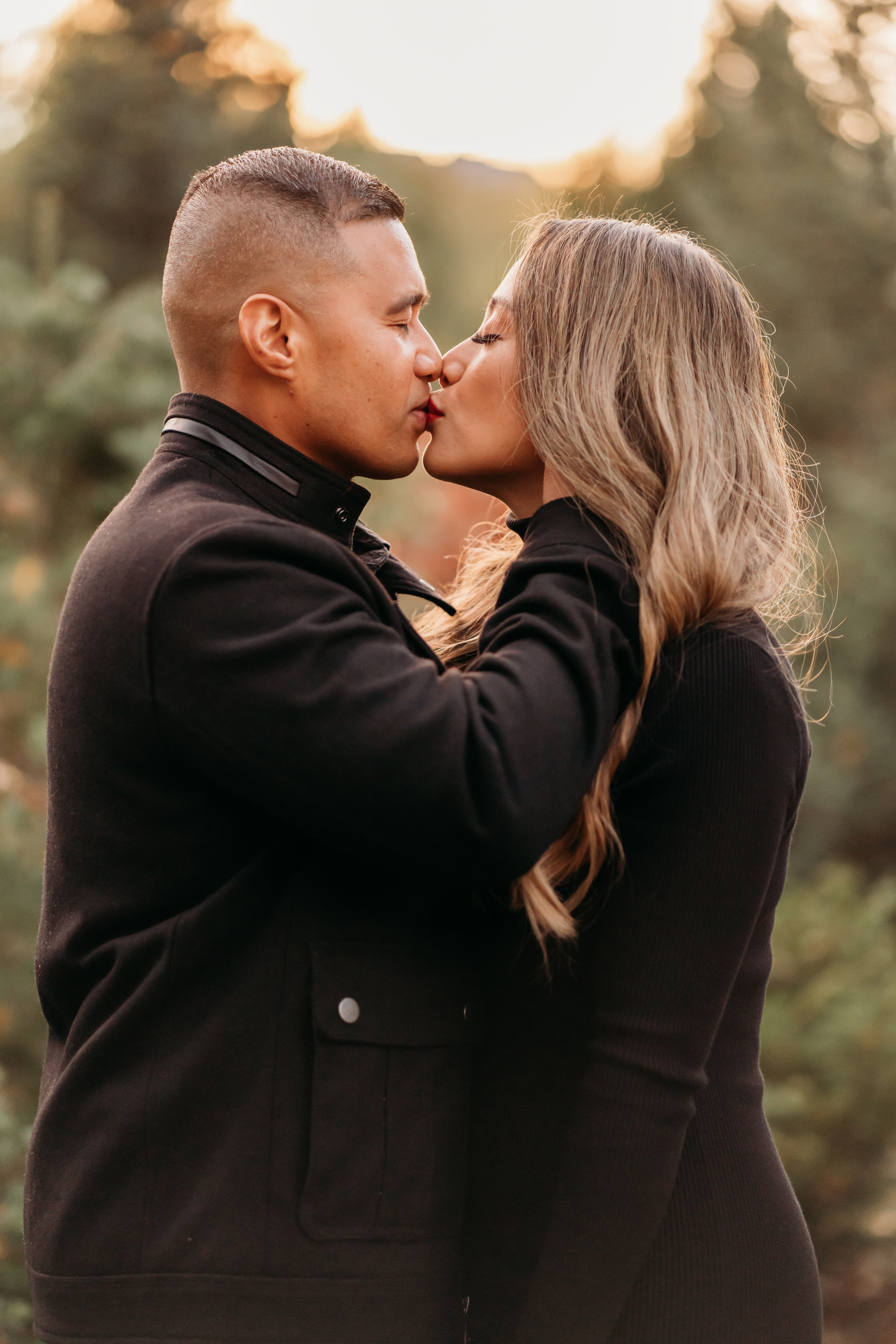 Couple in black jackets kiss nose to nose in a forest clearing at golden hour