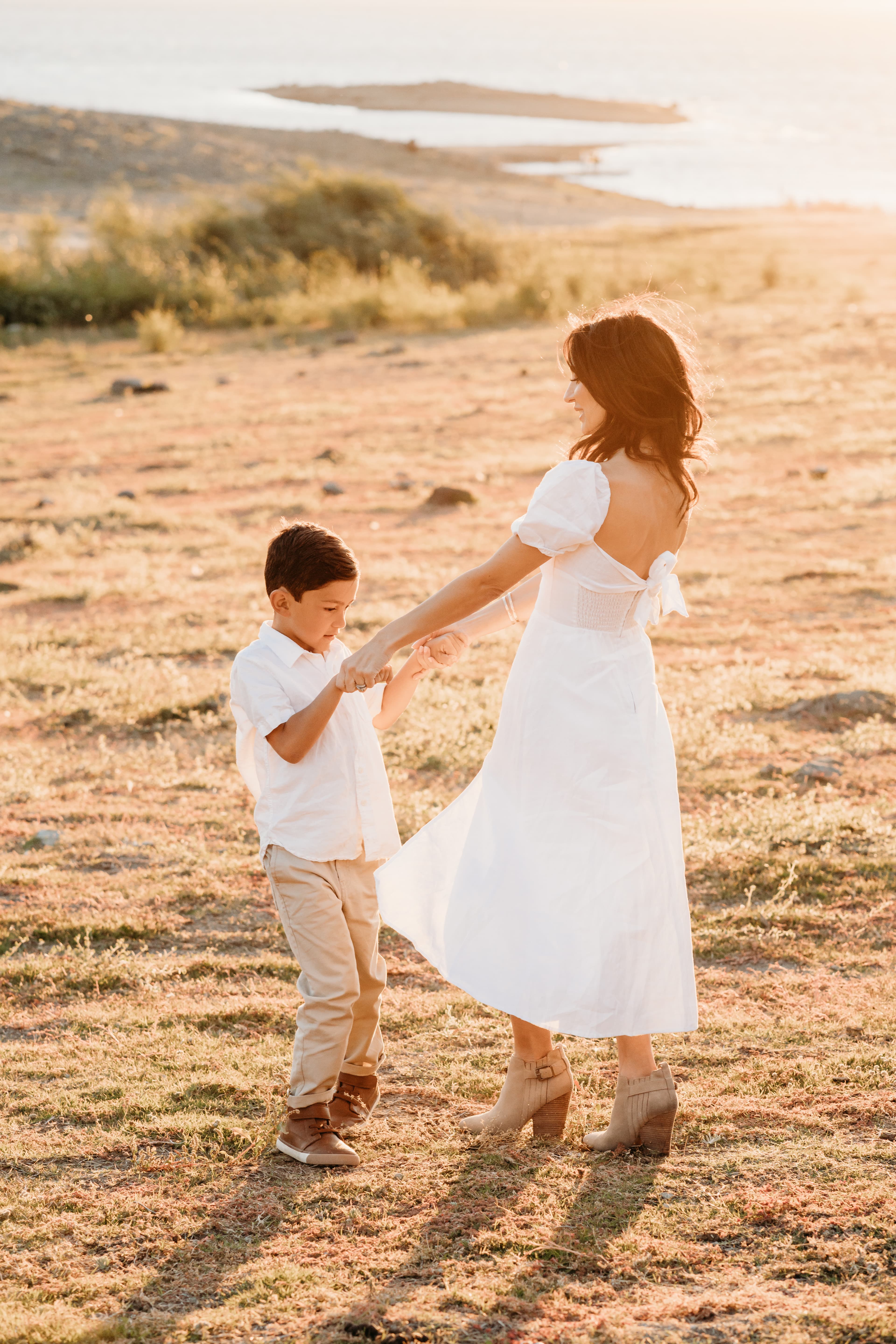 Mother in a white dress twirls her young son's hand in a golden sunset field