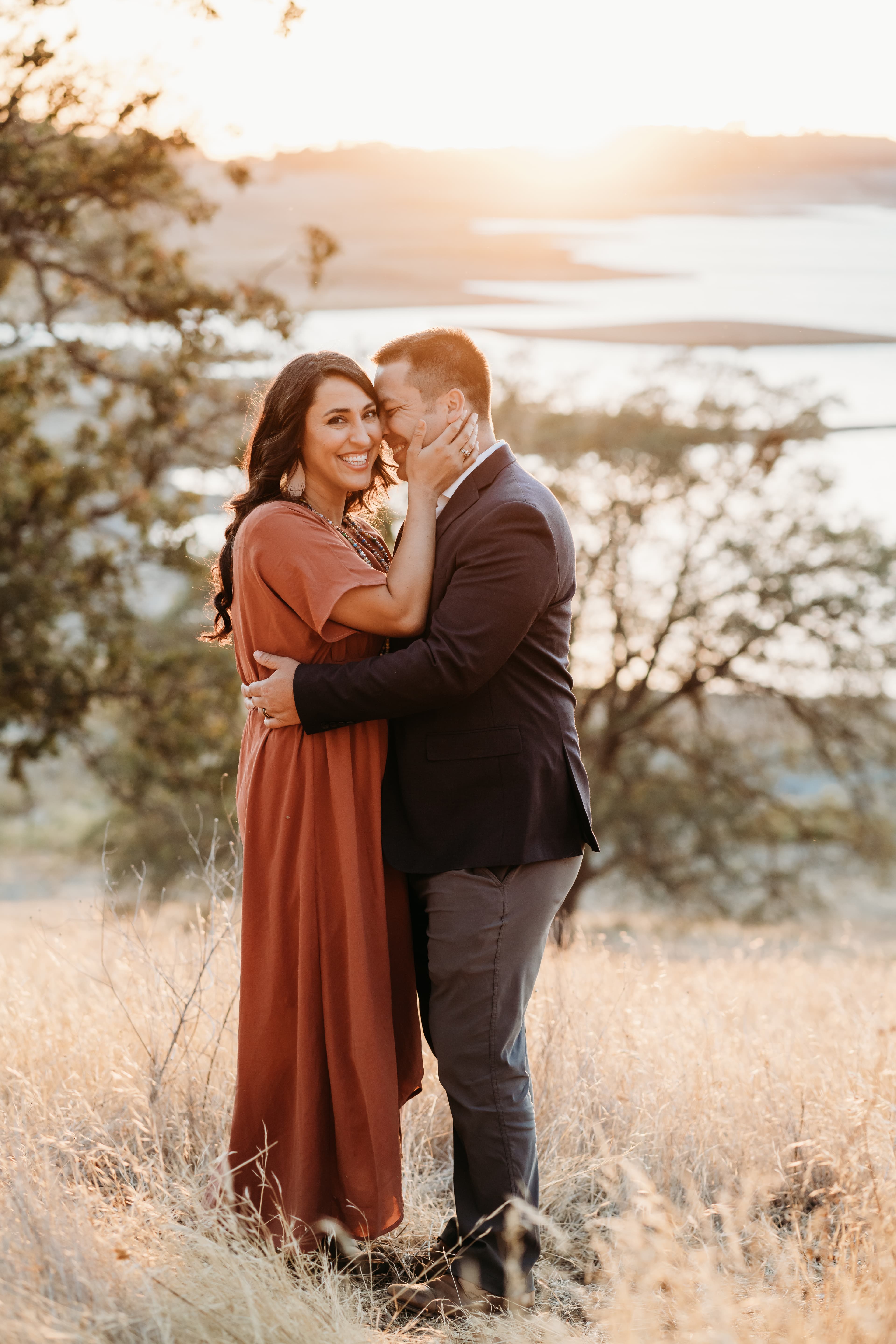 Couple in rust and navy embraces cheek to cheek overlooking a lake at sunset