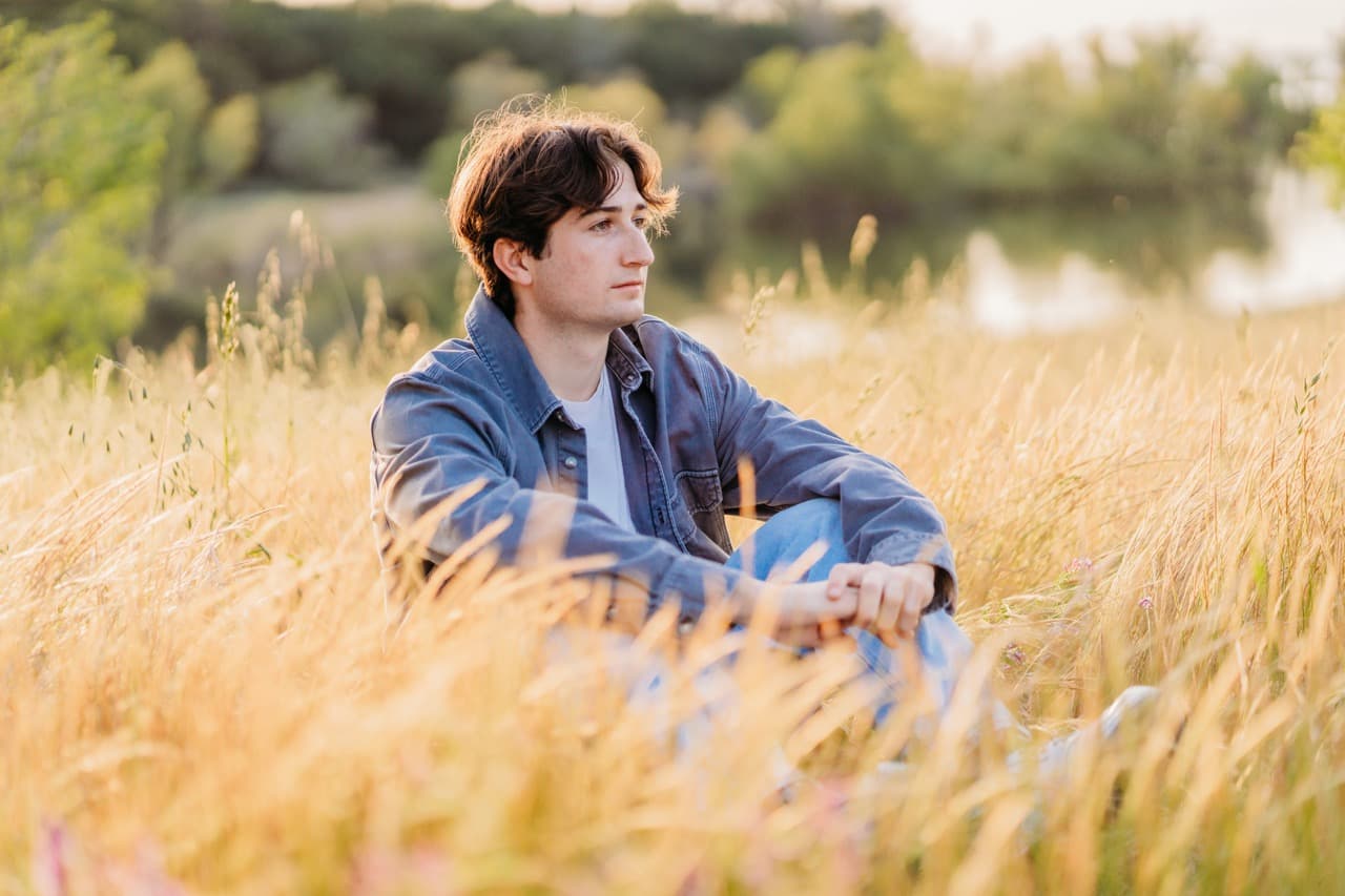 Young man in a denim jacket sits pensively in golden grass by a pond