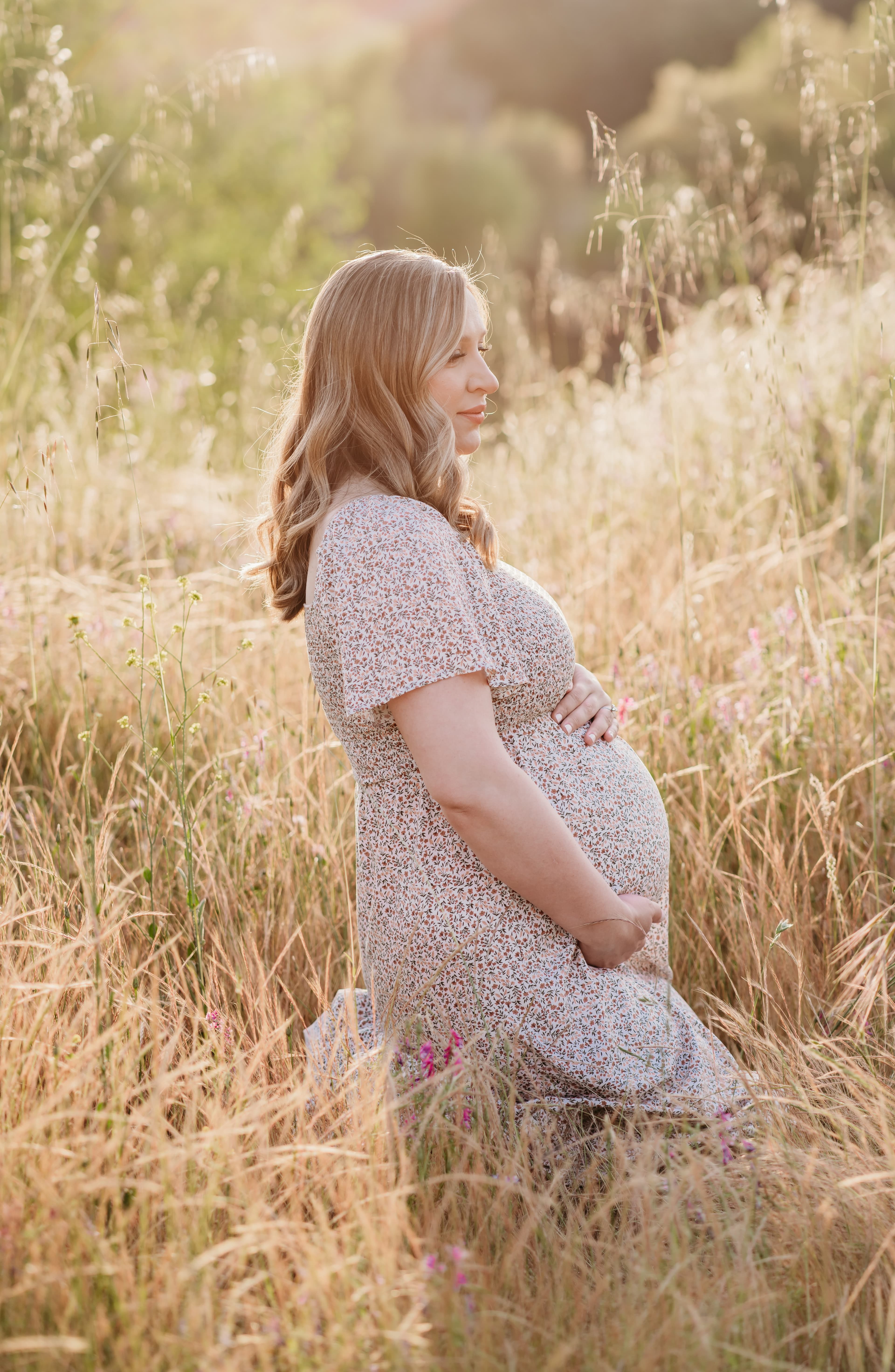 Pregnant woman in a floral dress cradles her belly while standing in a sunlit golden grass field