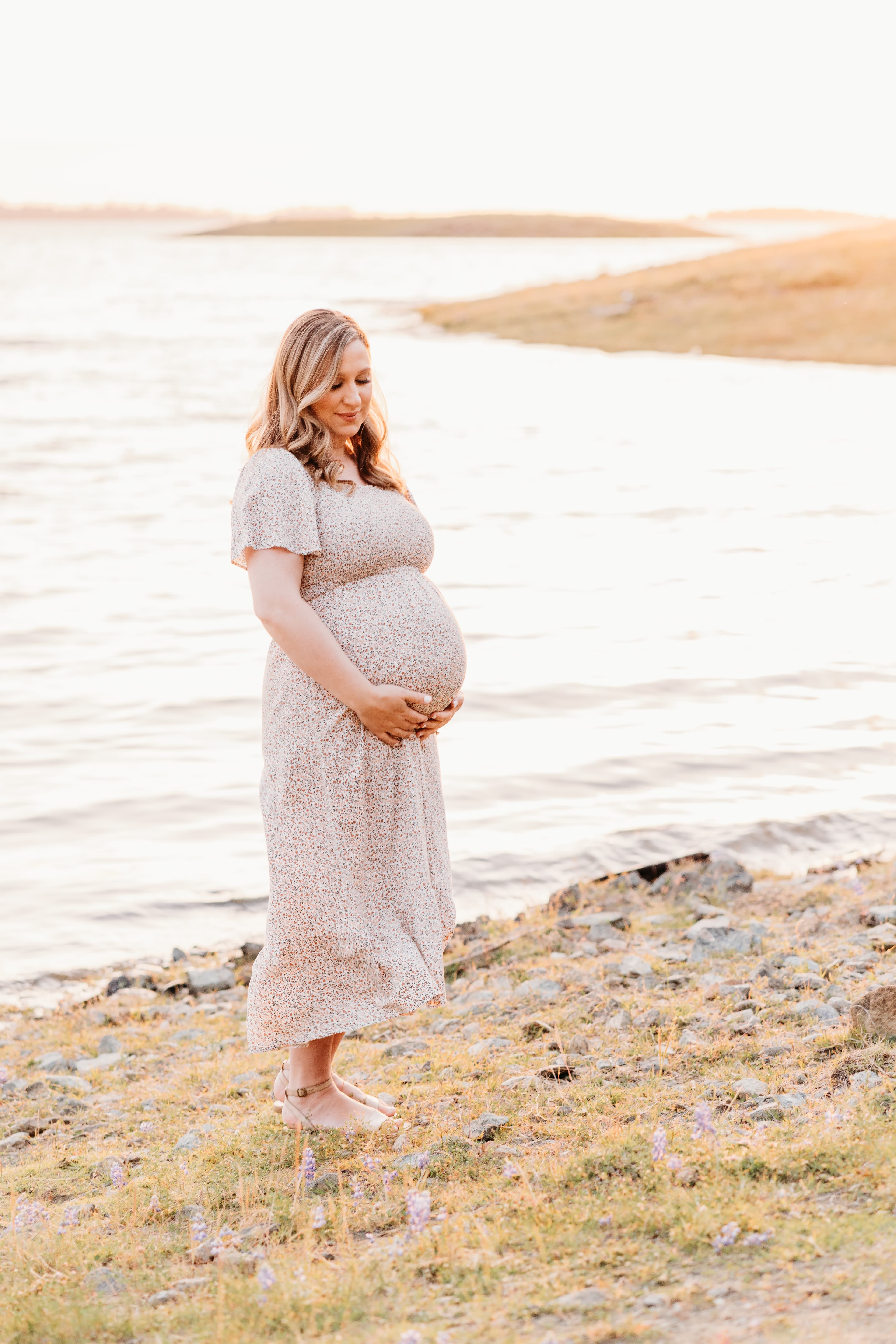 Pregnant woman in a floral dress stands at the water's edge, cradling her belly in golden sunset light
