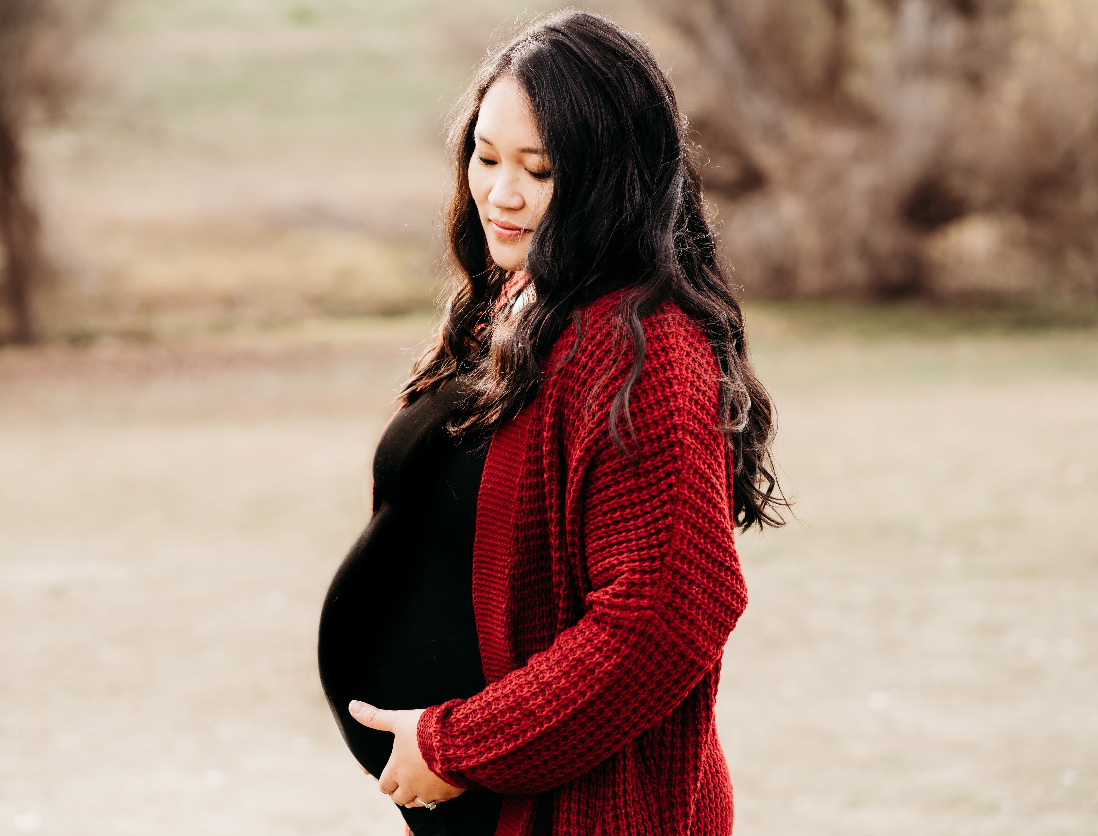 Pregnant woman in a red cardigan cradles her belly, looking down with a soft smile