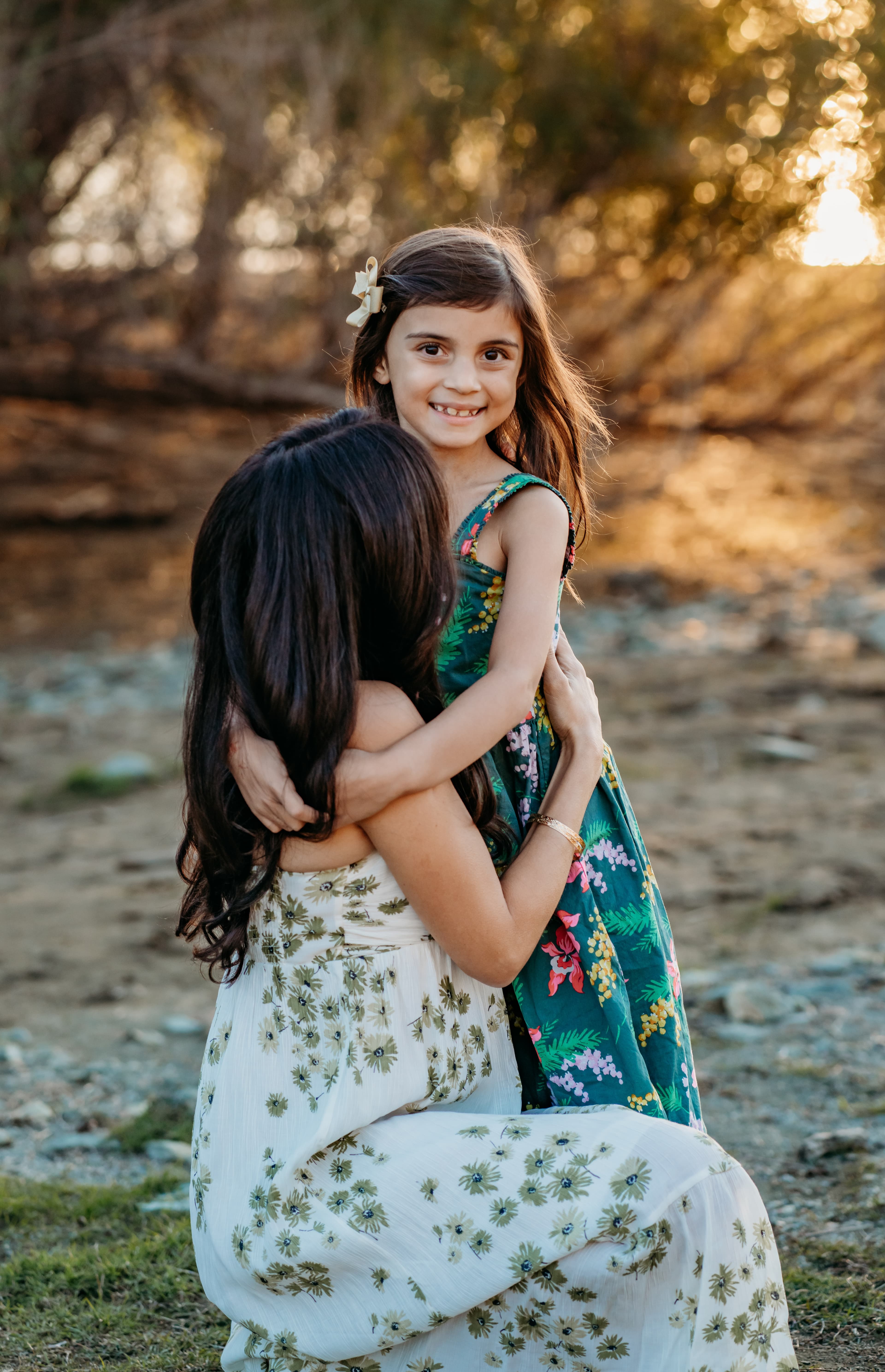 Mother kneels to hug her young daughter at golden hour by a creek