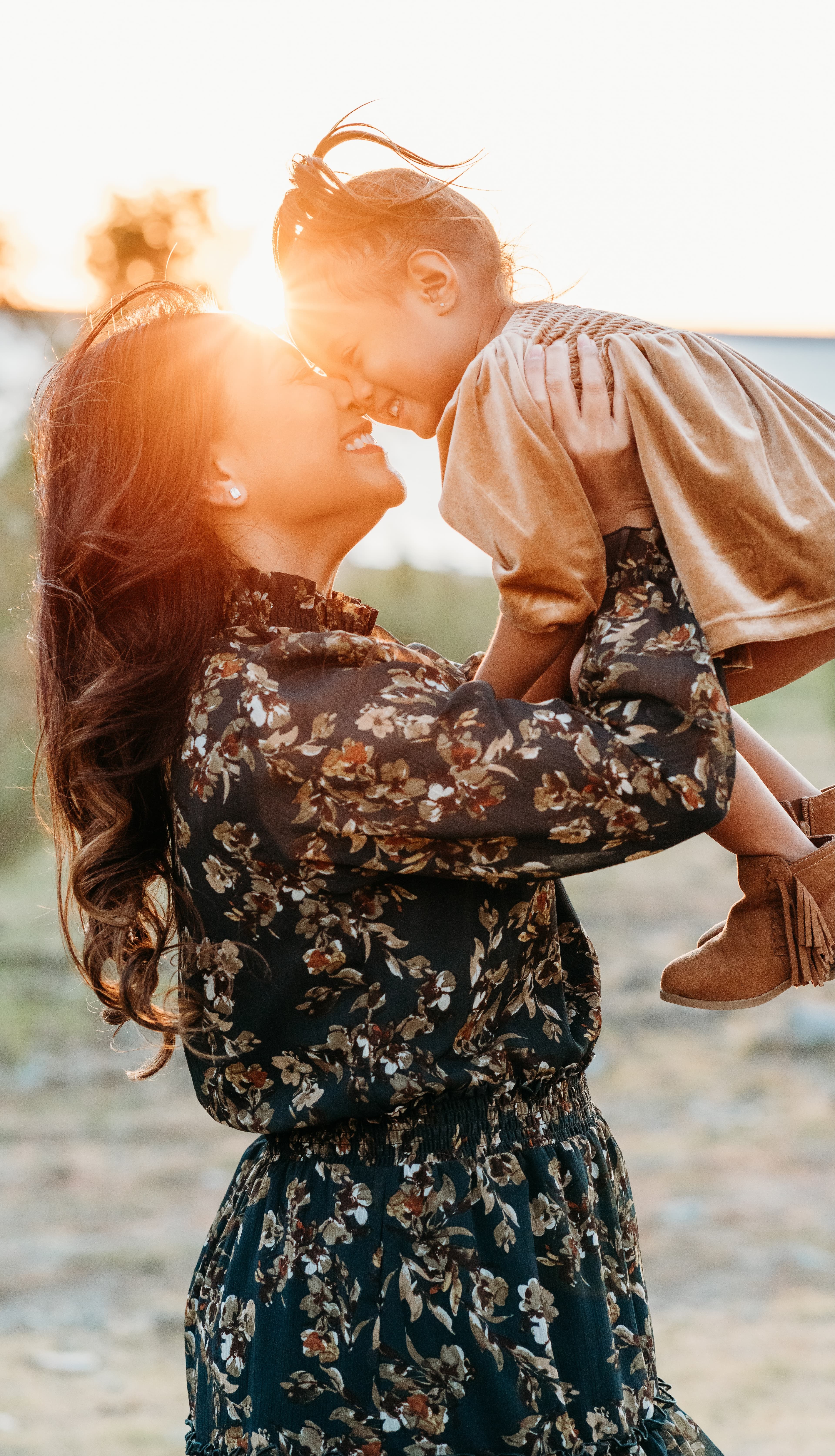 Mother lifts her young daughter nose to nose in front of a brilliant sunset flare