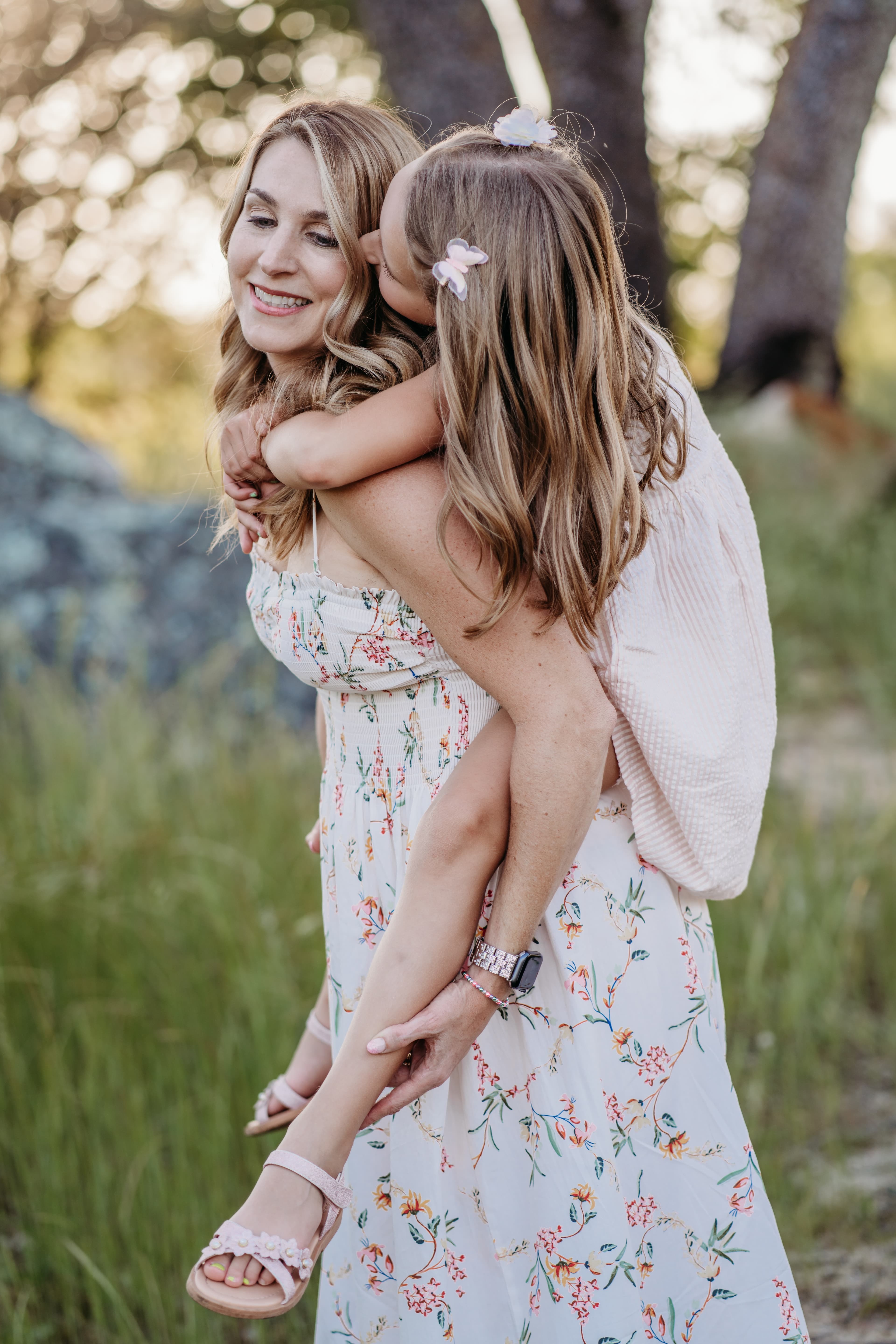 Mother carries her young daughter piggyback style, both smiling, in a sunlit outdoor setting