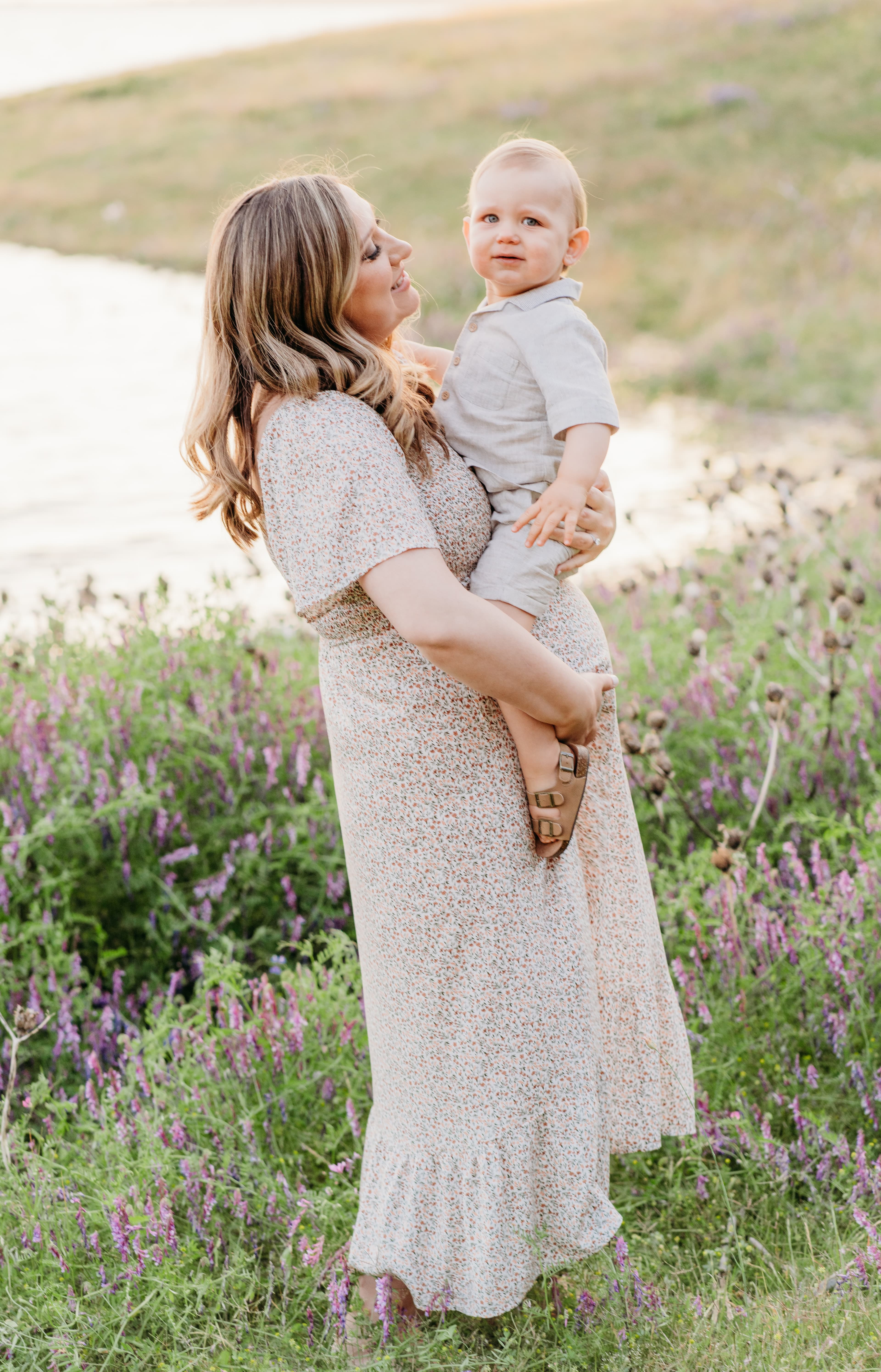 Mother in a floral dress holds her toddler son while standing in a wildflower field by the water