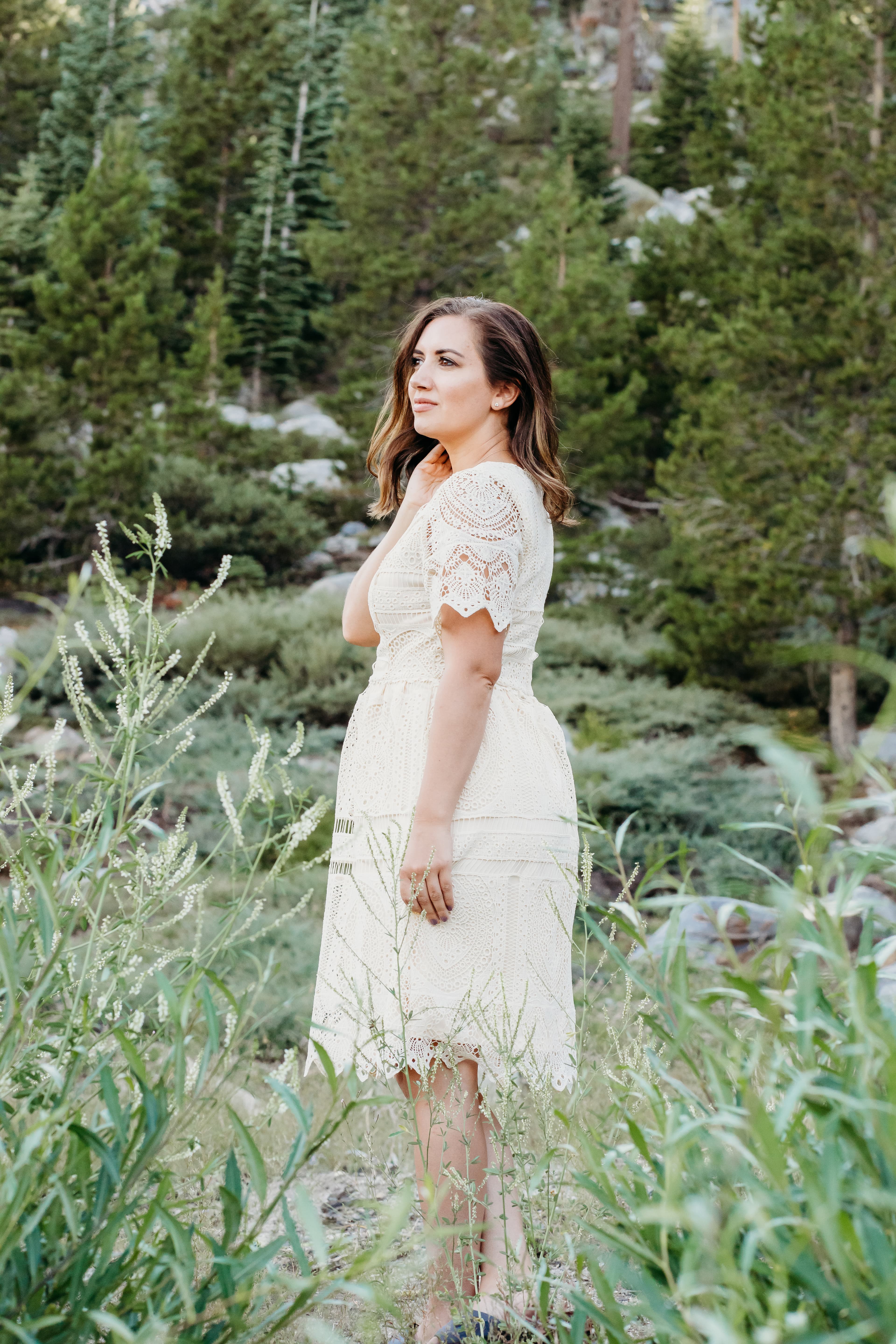 Woman in a white lace dress stands among pine trees, glancing to the side