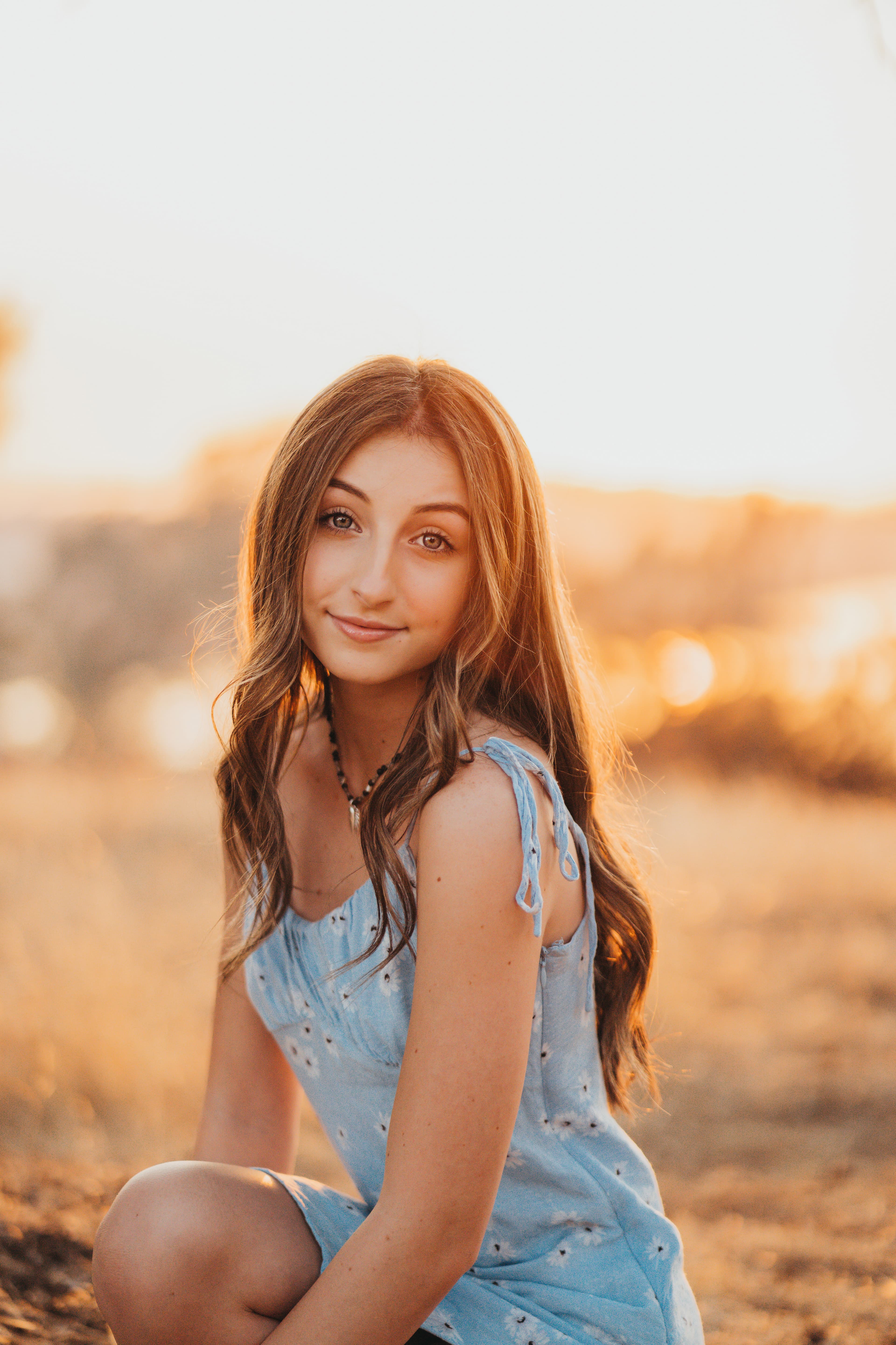 Teen girl in a blue floral dress crouches in golden grass, smiling