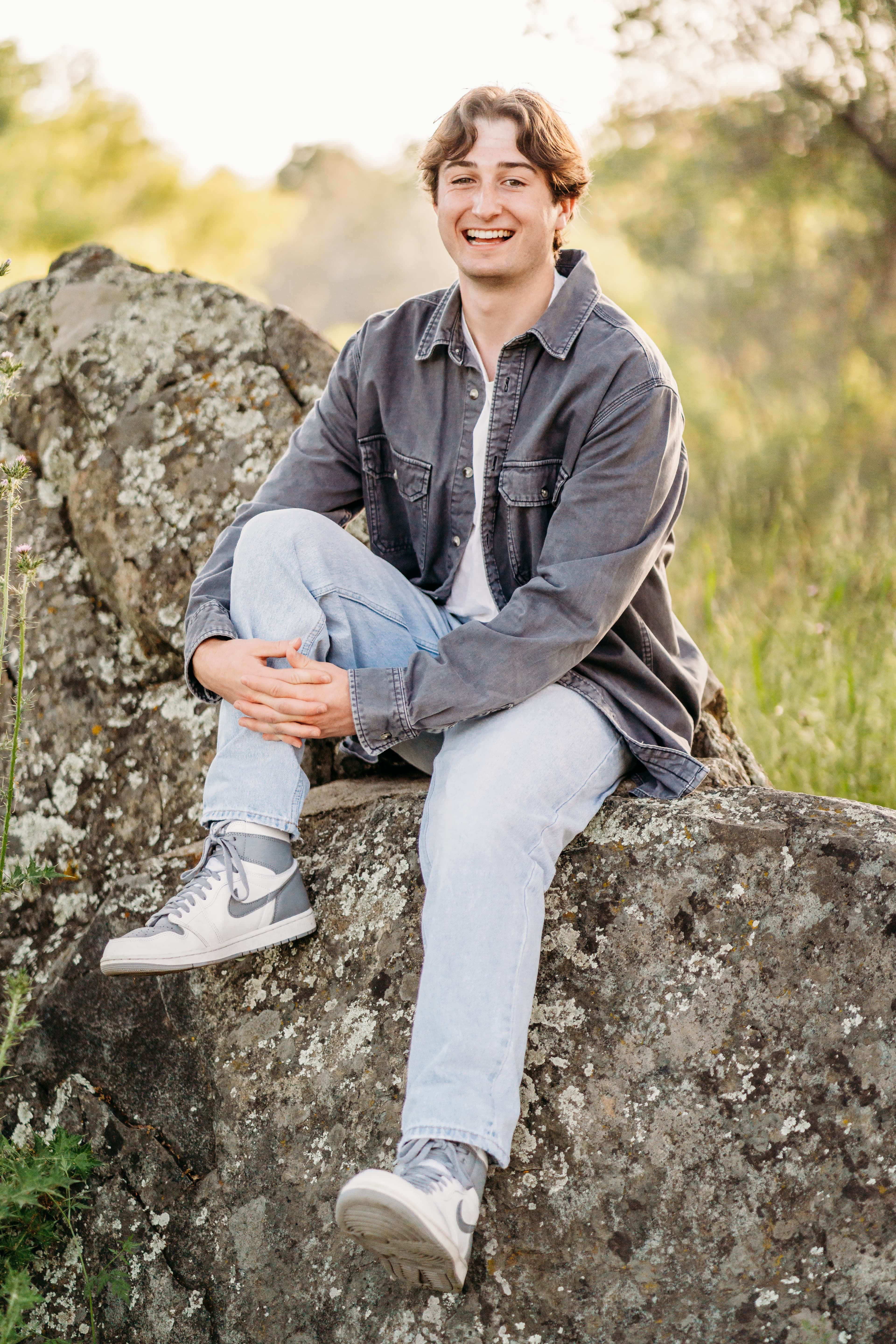 Teen boy in a denim shacket laughs on a wildflower-covered boulder