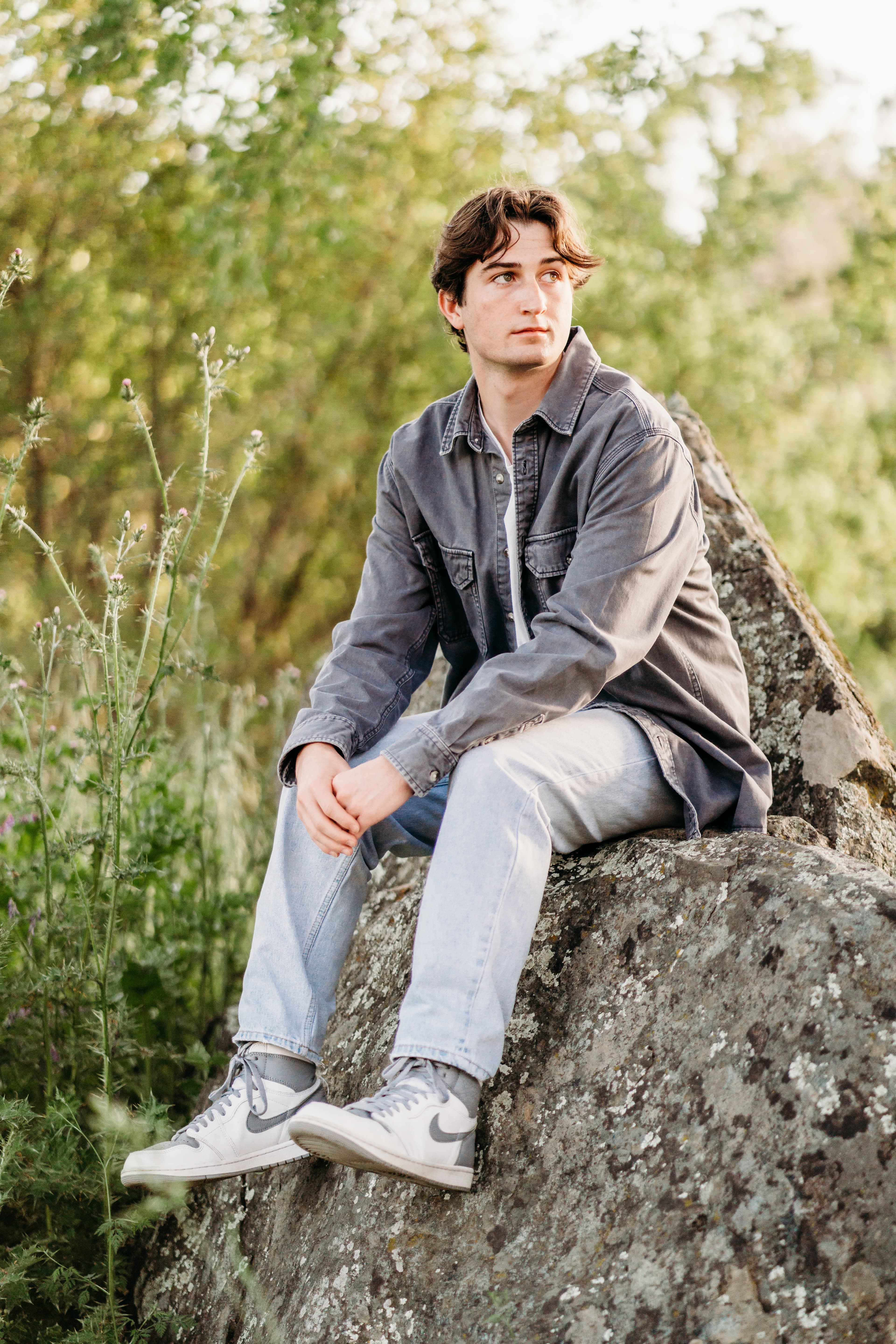 Teen boy in a denim shacket sits on a boulder among wildflowers, gazing off pensively