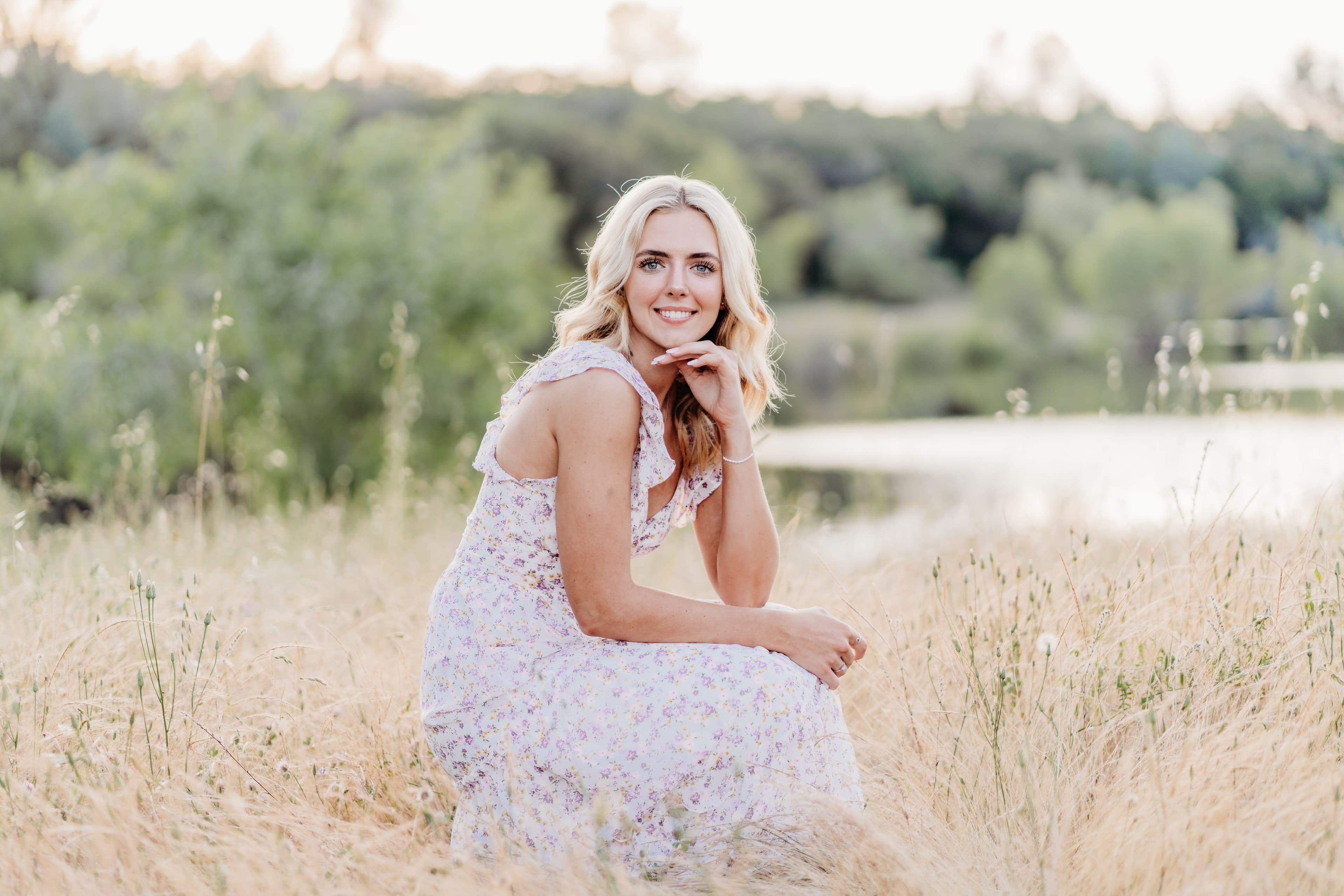 Young woman crouches with her chin resting on her hand near a lake, soft smile