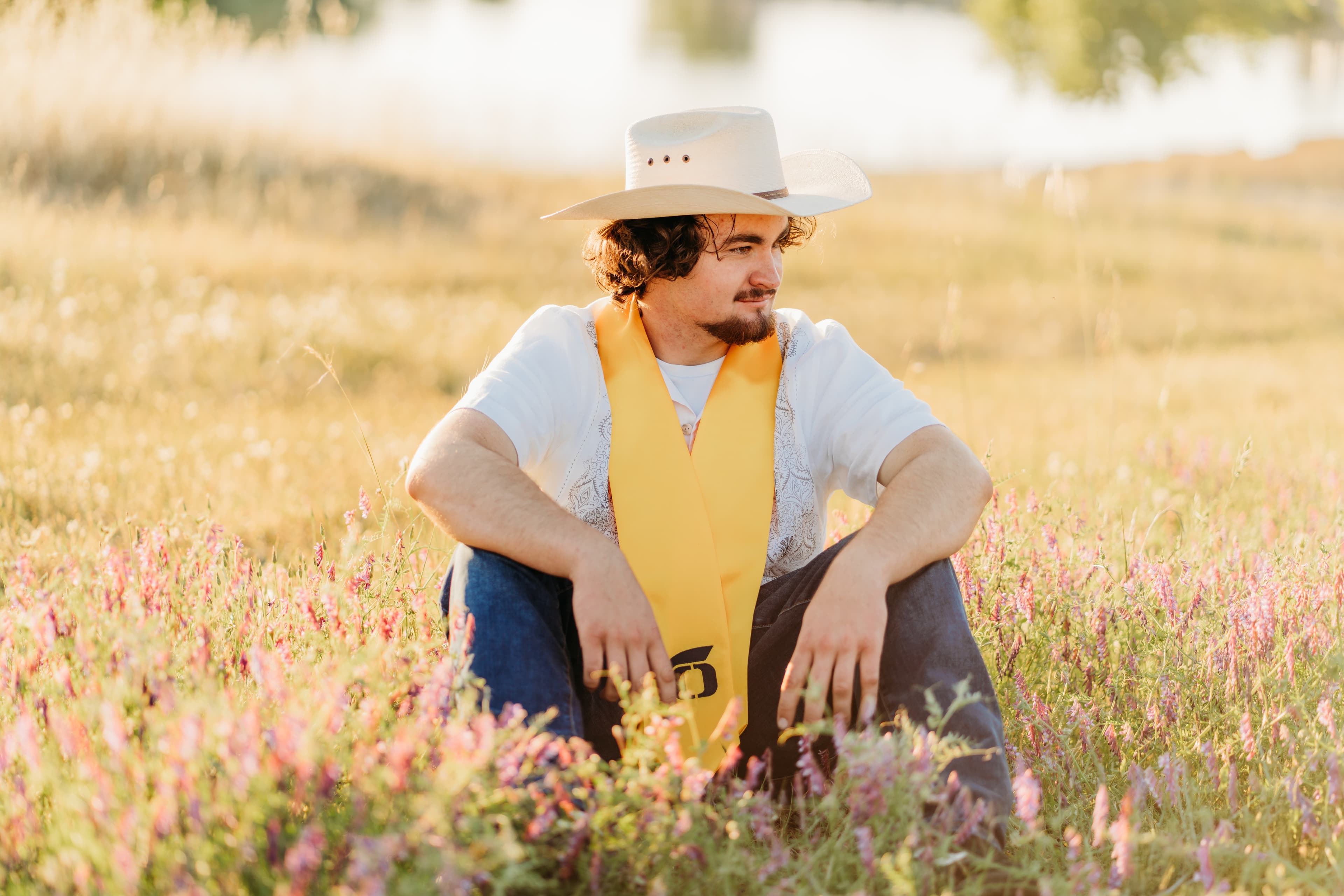 Graduate in a cowboy hat and yellow stole sits in a wildflower field by a lake