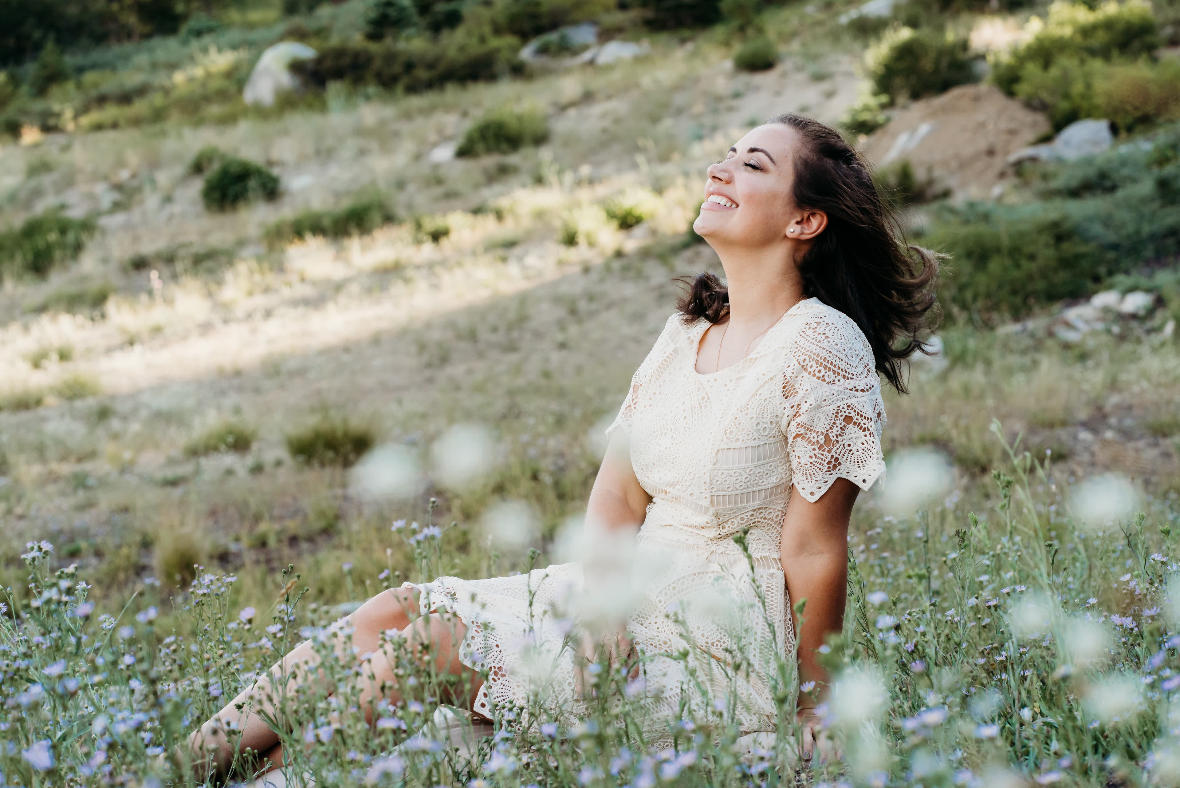 Young woman in a cream lace dress leans back laughing in a mountain meadow