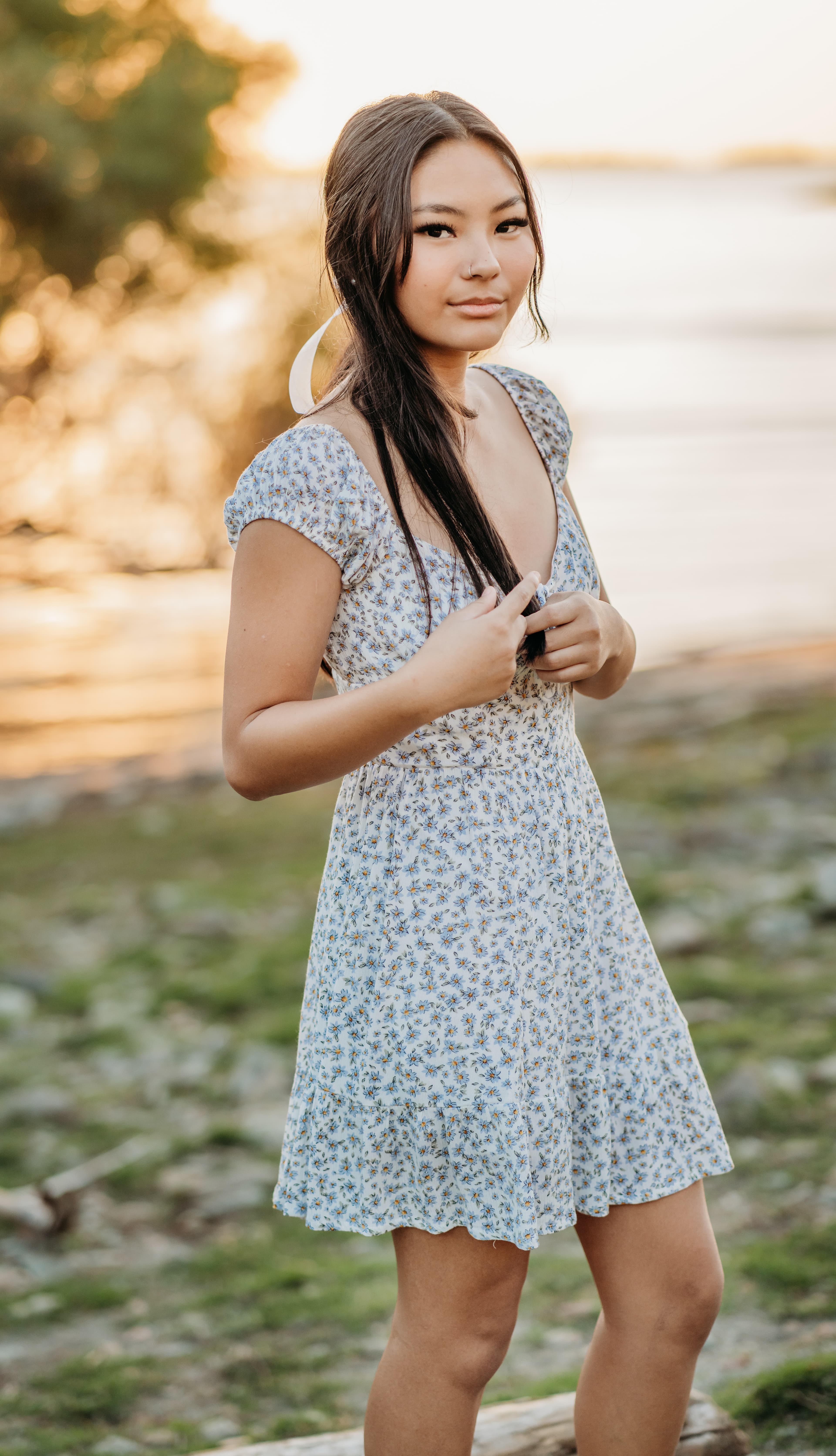 Young woman in a floral dress stands on a log by the water, smiling softly at the camera