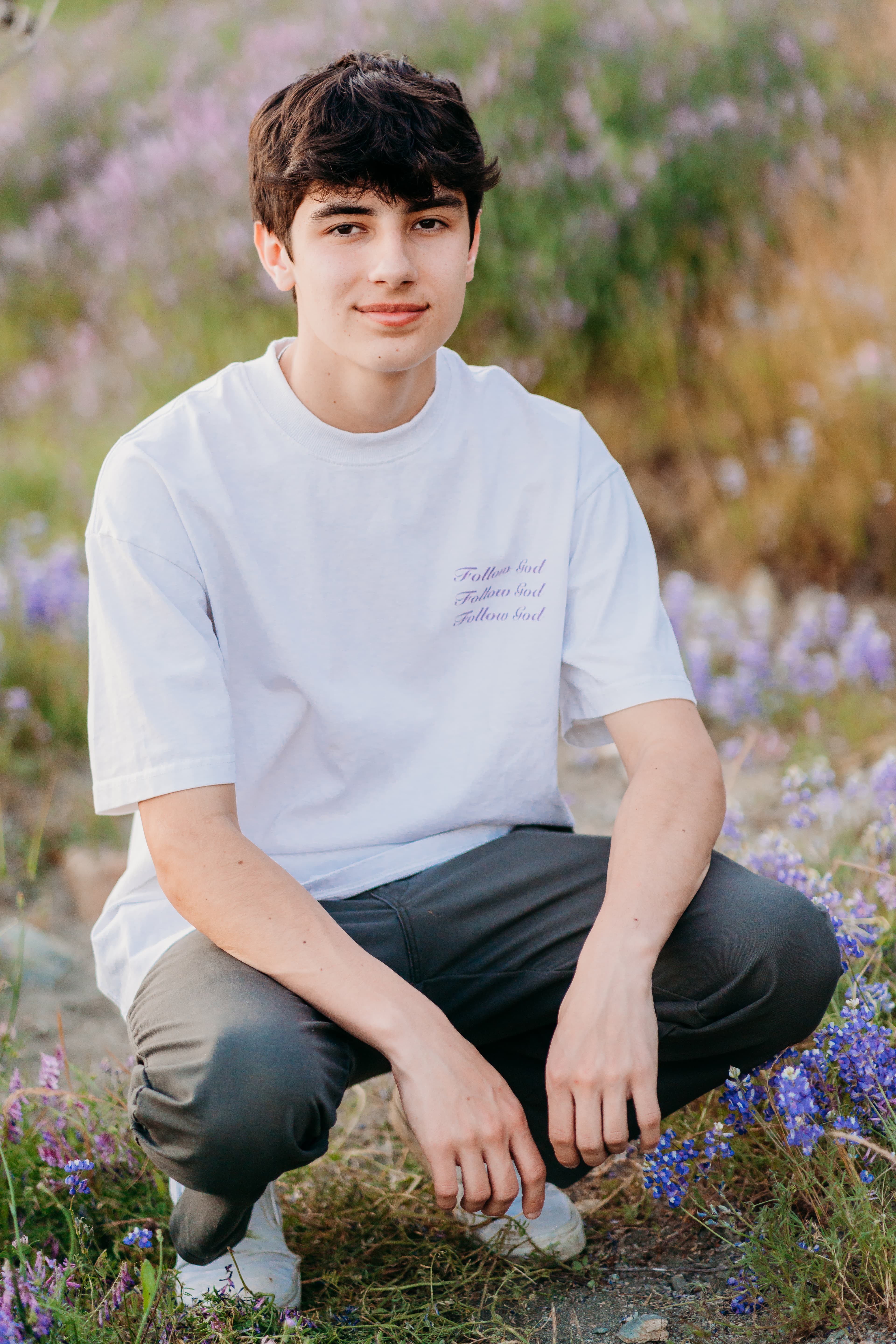 Teen boy crouches in a wildflower field wearing a white graphic tee